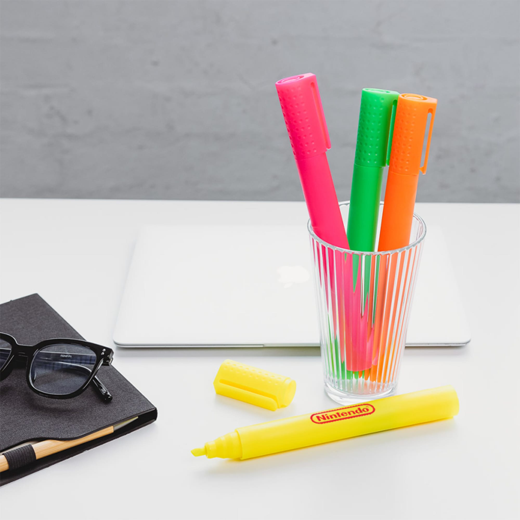 A glass holding pink, green, and orange jumbo highlighters alongside a yellow highlighter, with a notebook and glasses nearby.