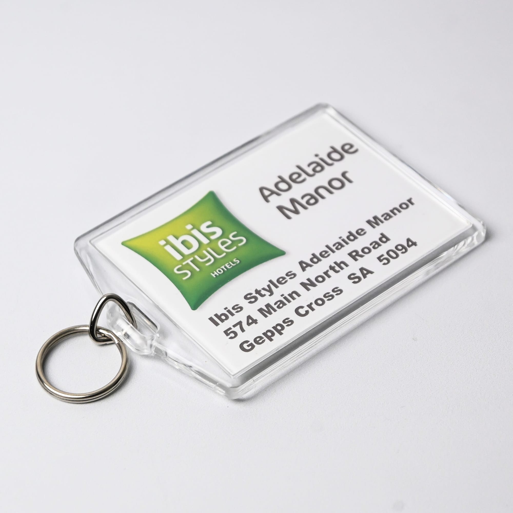 A clear hotel keychain with a ring, featuring green and white branding and a printed address. Australian made.