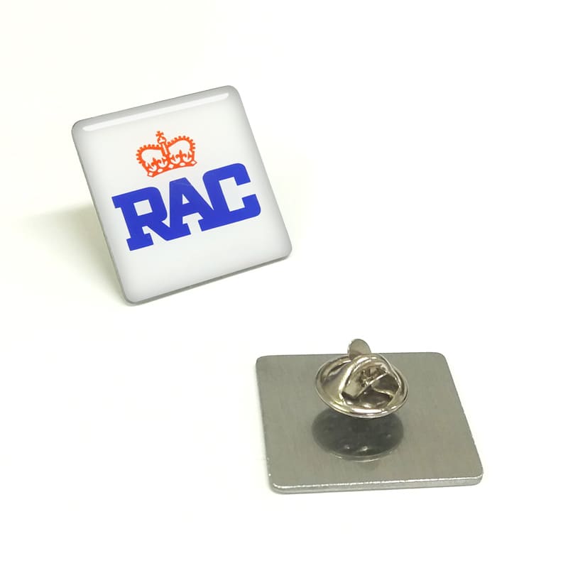 A square lapel pin featuring a white background with blue lettering and a logo, along with its metal backing.