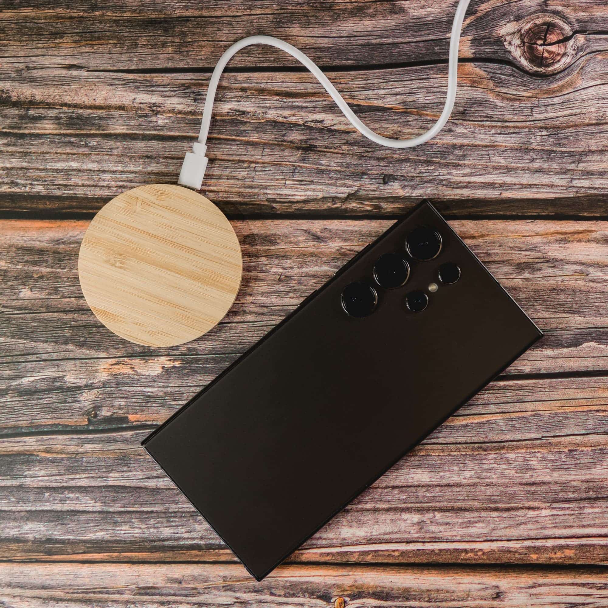 A round bamboo wireless charger alongside a smartphone, connected with a charging cable.