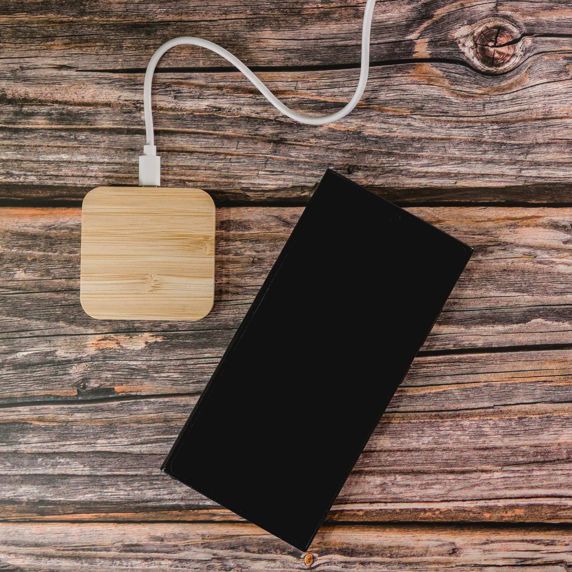 A bamboo wireless charger with a white charging cable is placed next to a black smartphone on a wooden surface.