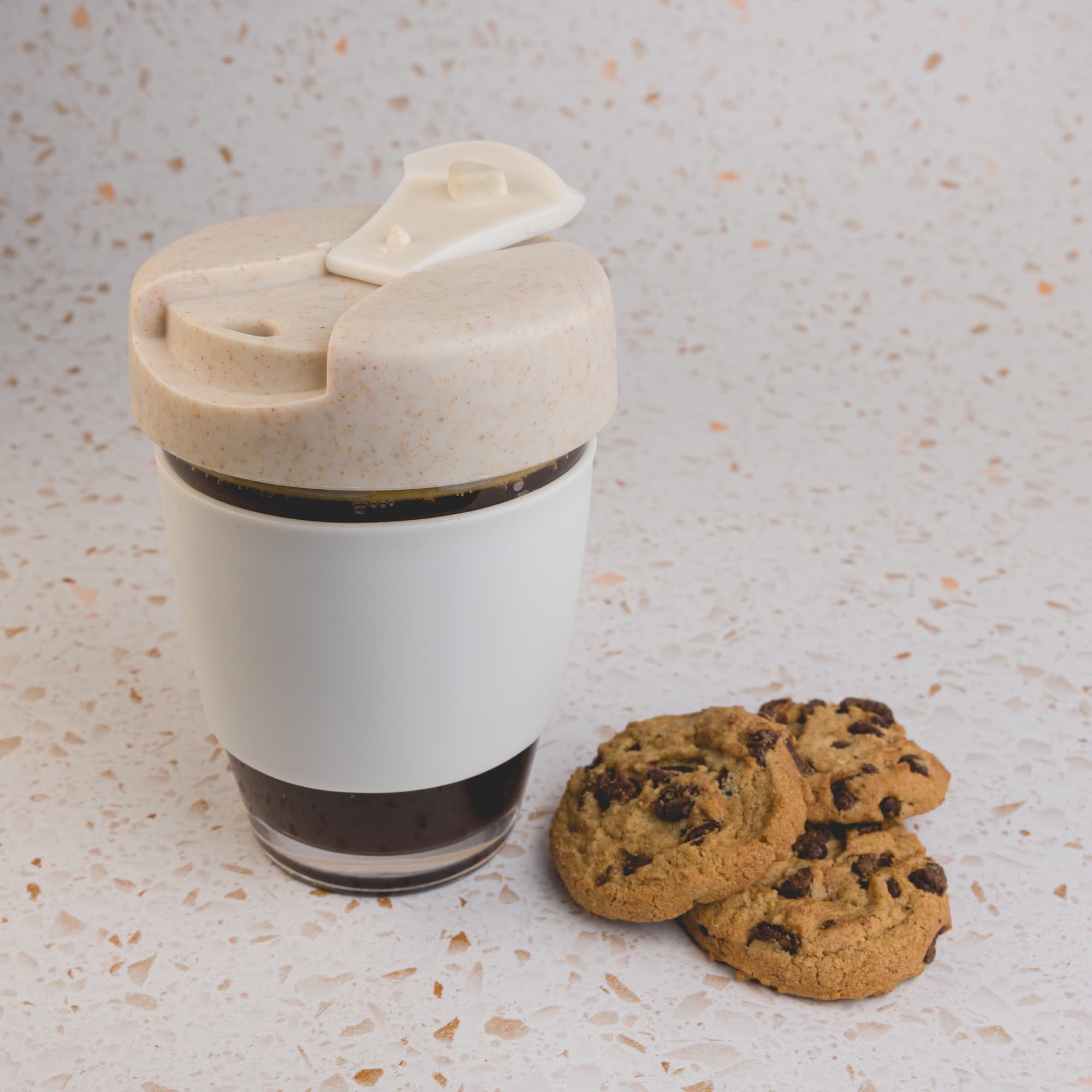 A reusable coffee cup with a beige lid and white base, alongside three chocolate chip cookies on a speckled surface.