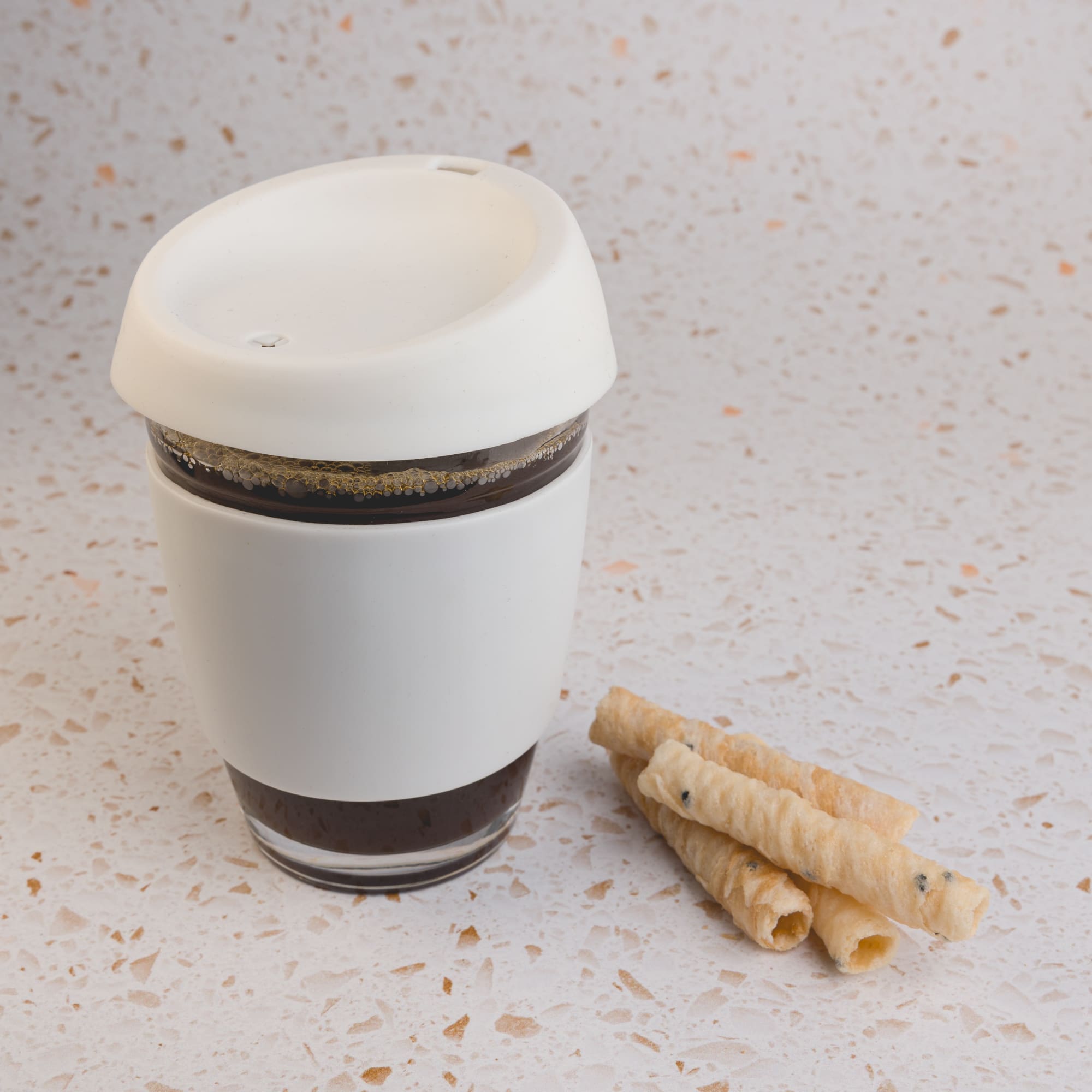 A reusable coffee cup with a silicone lid, featuring a sleek design in white, accompanied by rolled wafers.