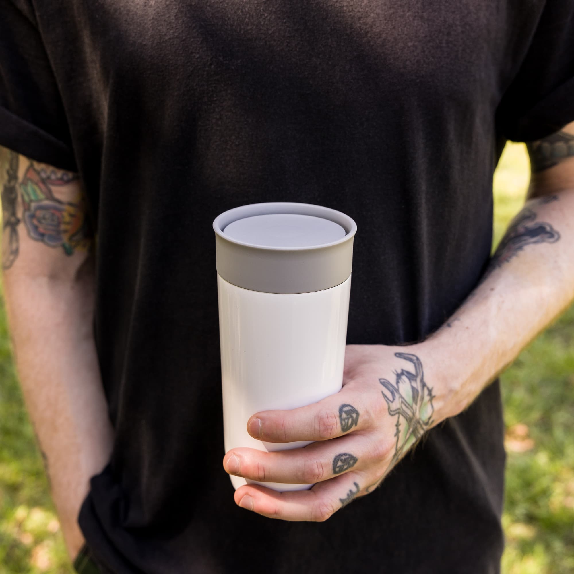 A reusable stainless steel coffee cup with a white body and gray lid, held by a person with tattoos.