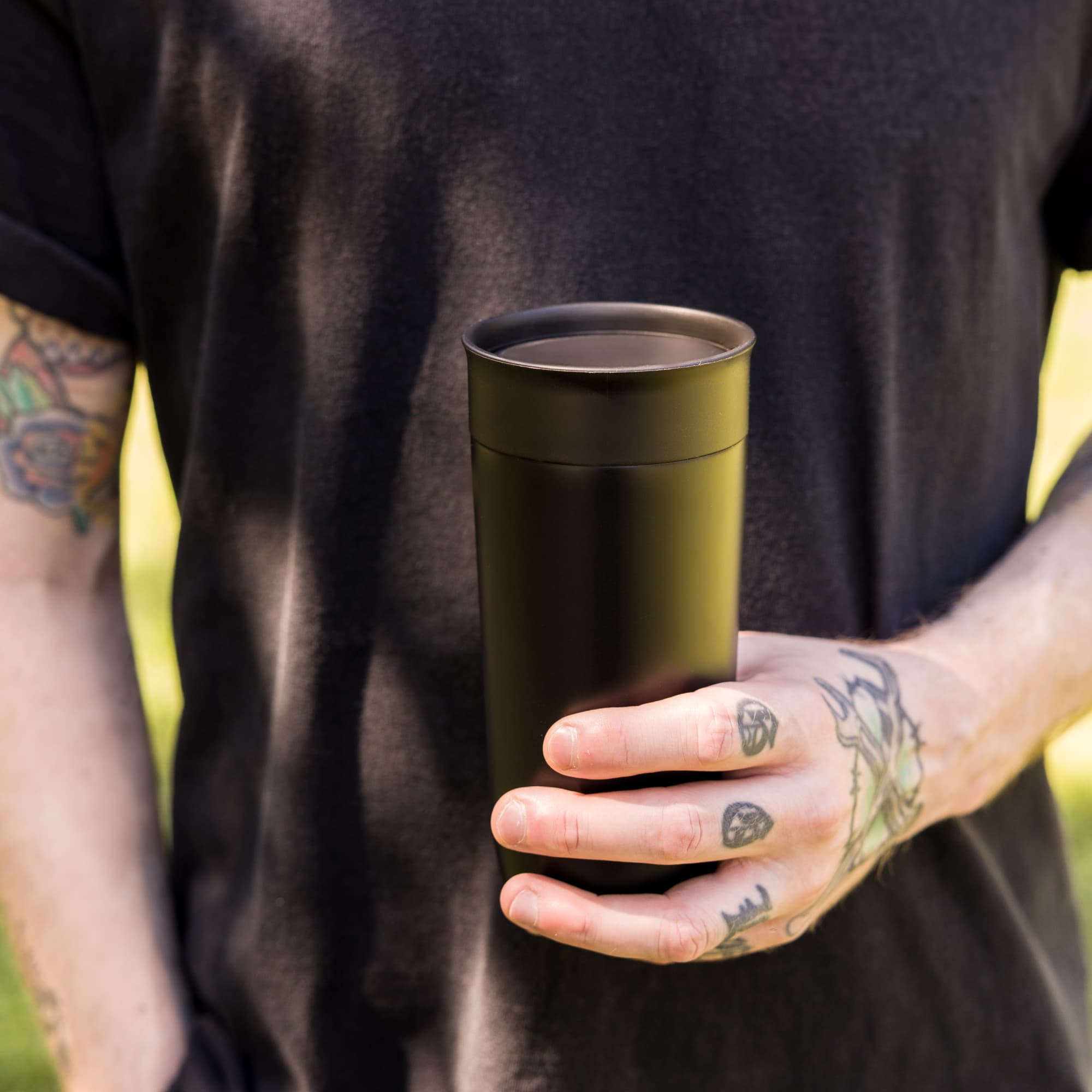 A stainless steel coffee cup with a sleek black finish, held by a person with tattoos.