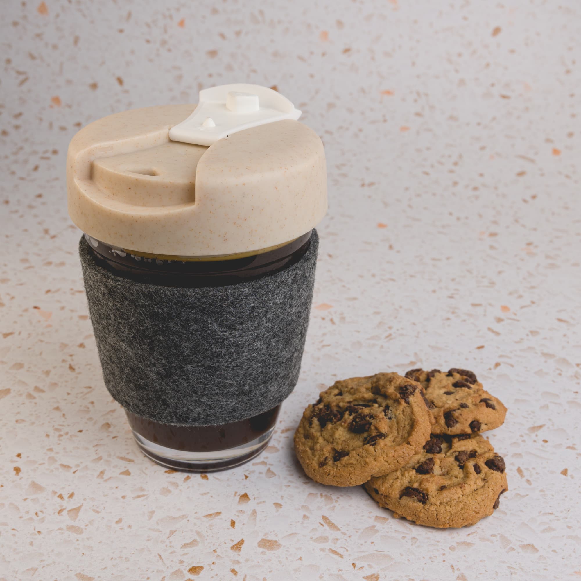 A reusable coffee cup with a beige lid and a dark grey RPET band, accompanied by a few chocolate chip cookies.