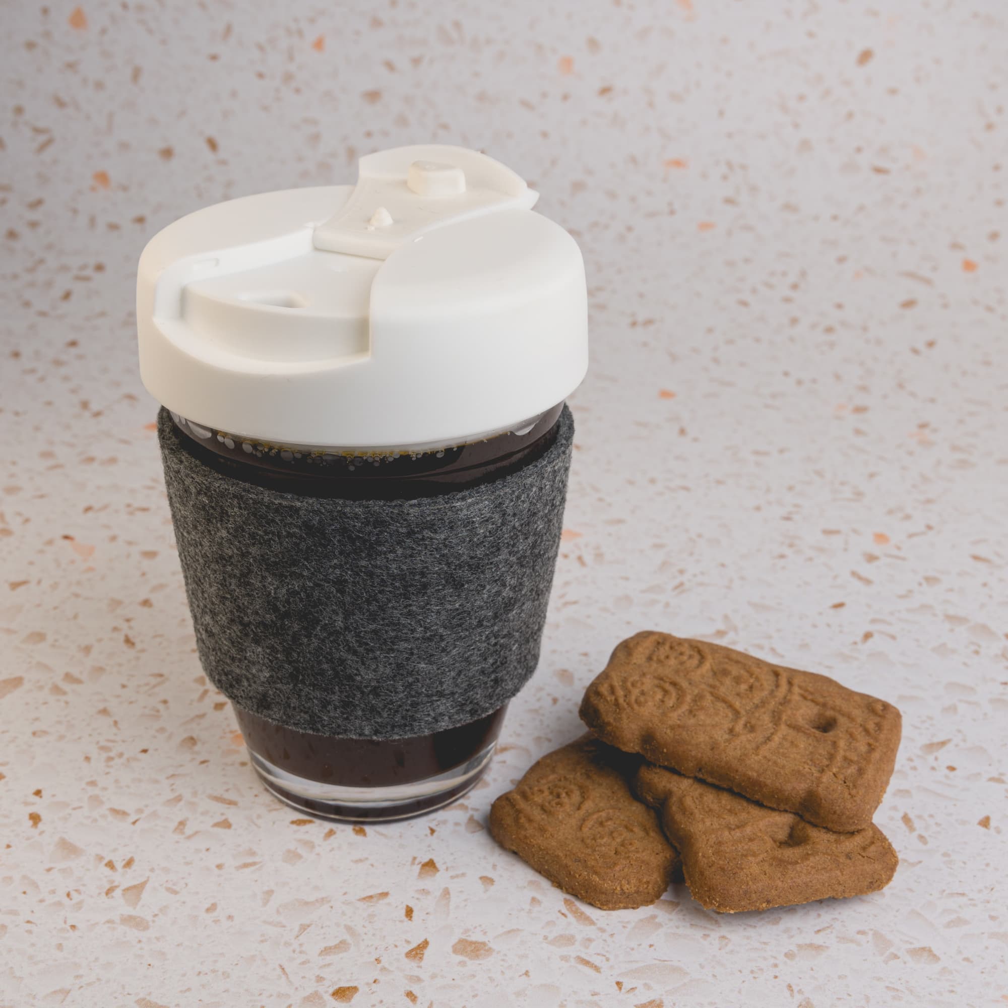 A reusable coffee cup with a flip lid and a felt RPET band is placed next to a few biscuits.