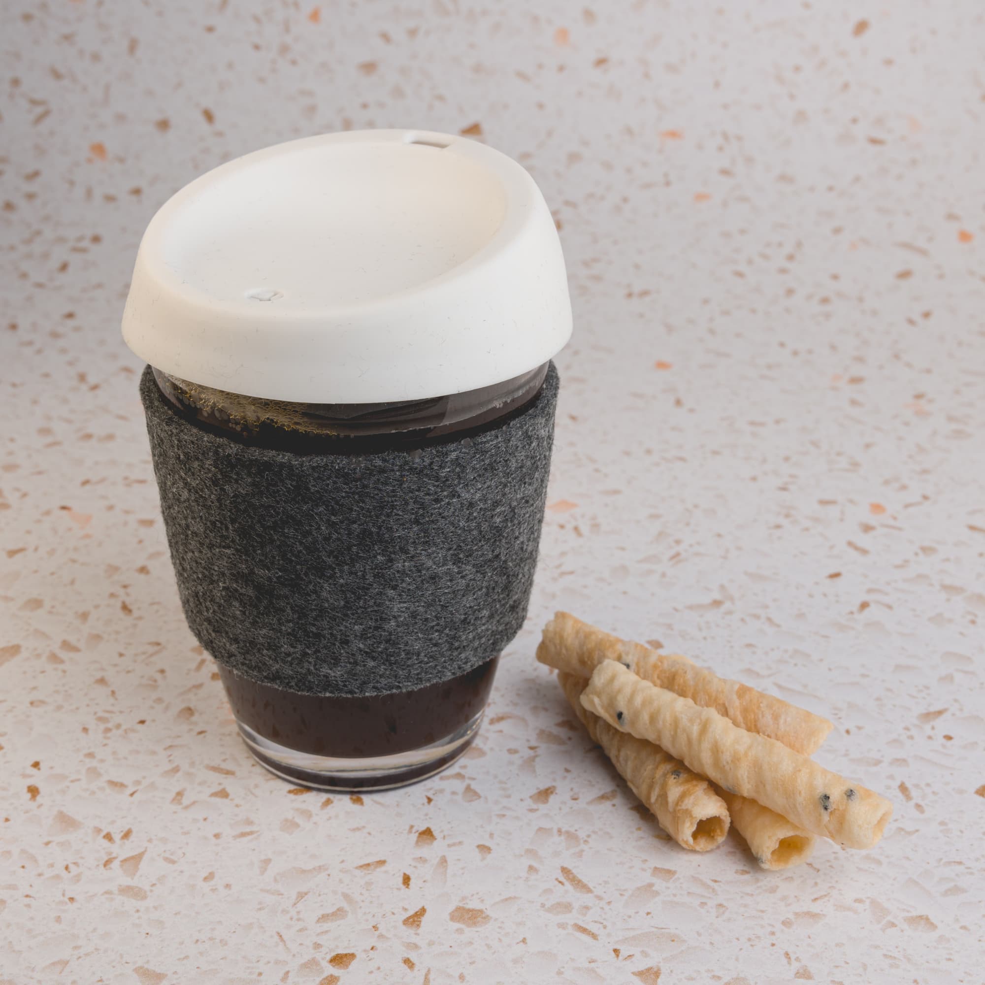 A reusable coffee cup featuring a silicone lid and a RPET band, accompanied by some rolled wafers.