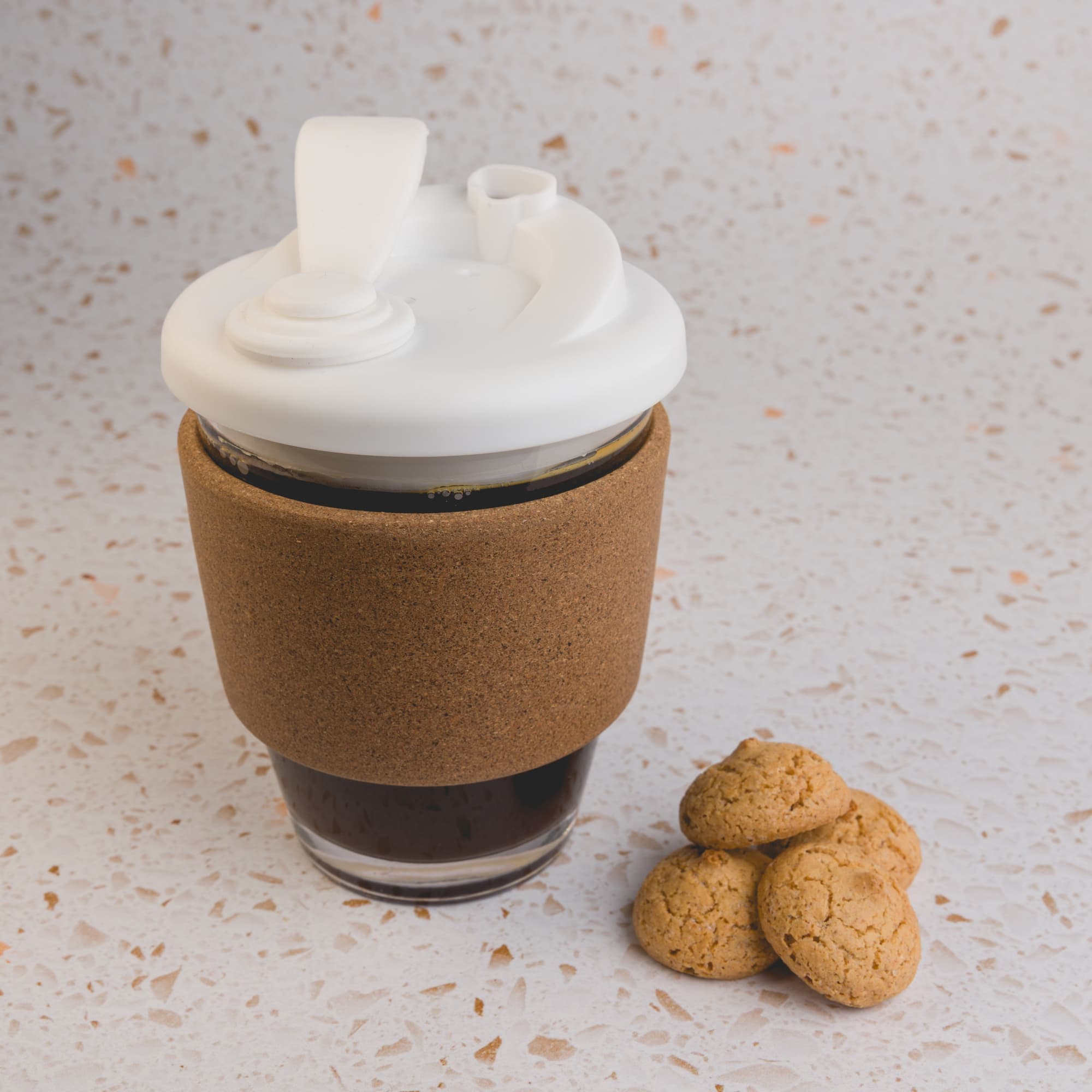 A reusable coffee cup featuring a white snap lid and a cork band, accompanied by a few cookies.