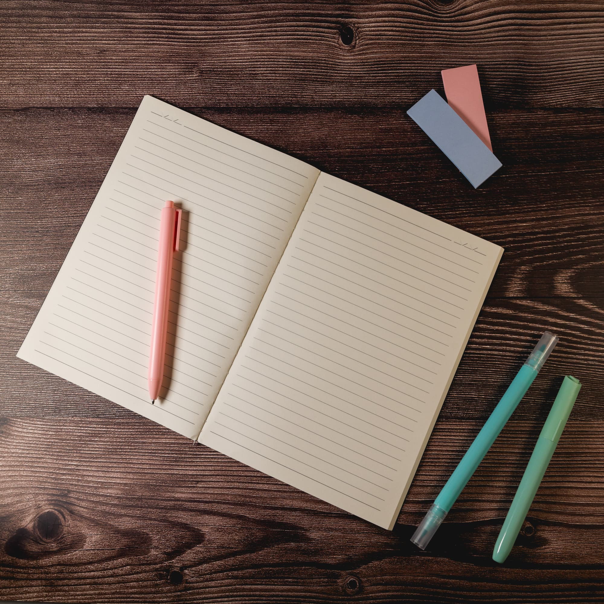A light-colored notebook with lined pages sits on a wooden surface, accompanied by a pink pen and colored highlighters.