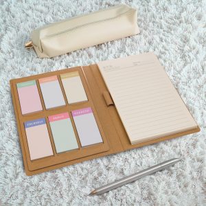 A planner in a tan folder with multiple sticky note pads in pastel colors and a pen beside a cream pouch.