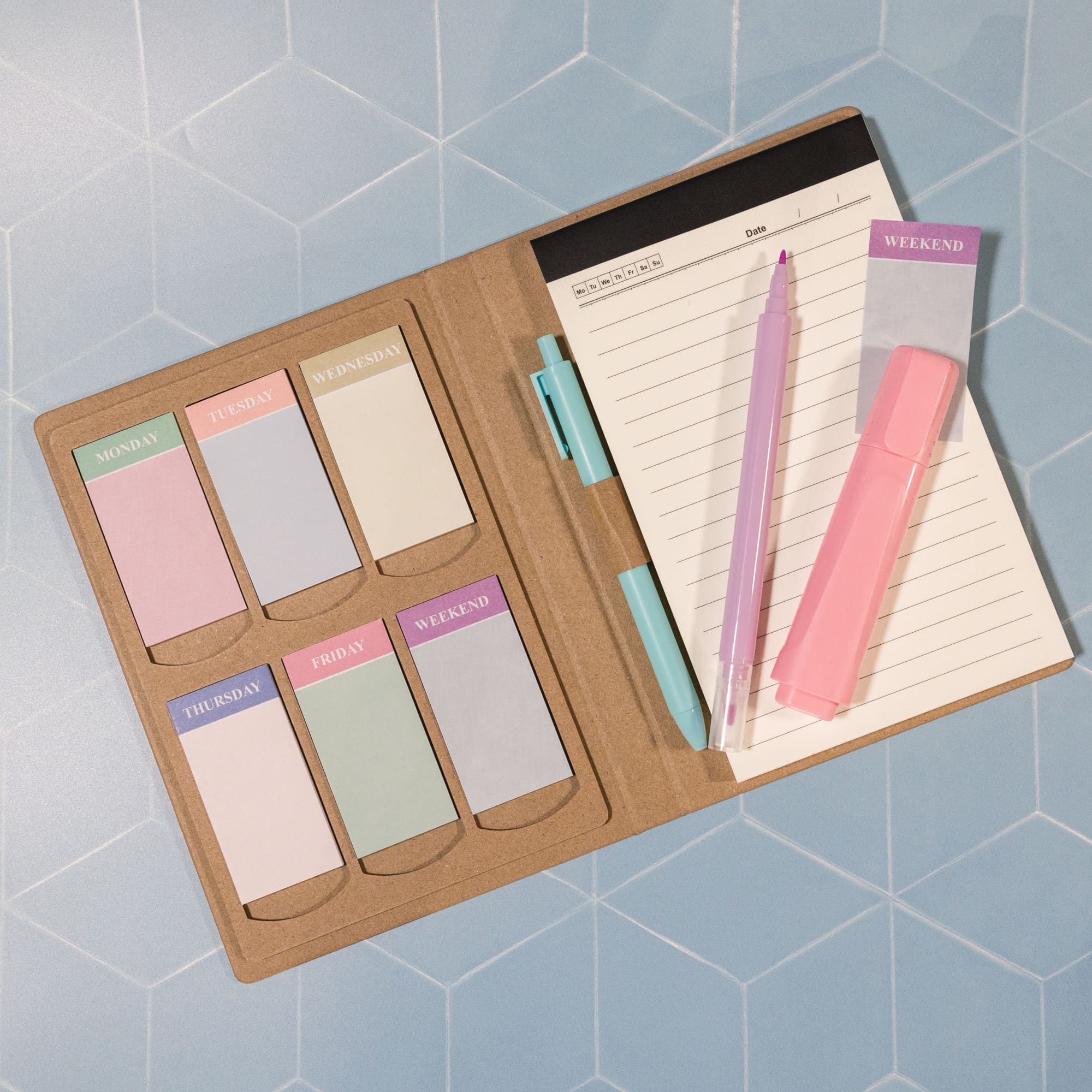 A planner with a tan cover, featuring colorful tabs for each weekday and pastel pink and purple accessories.