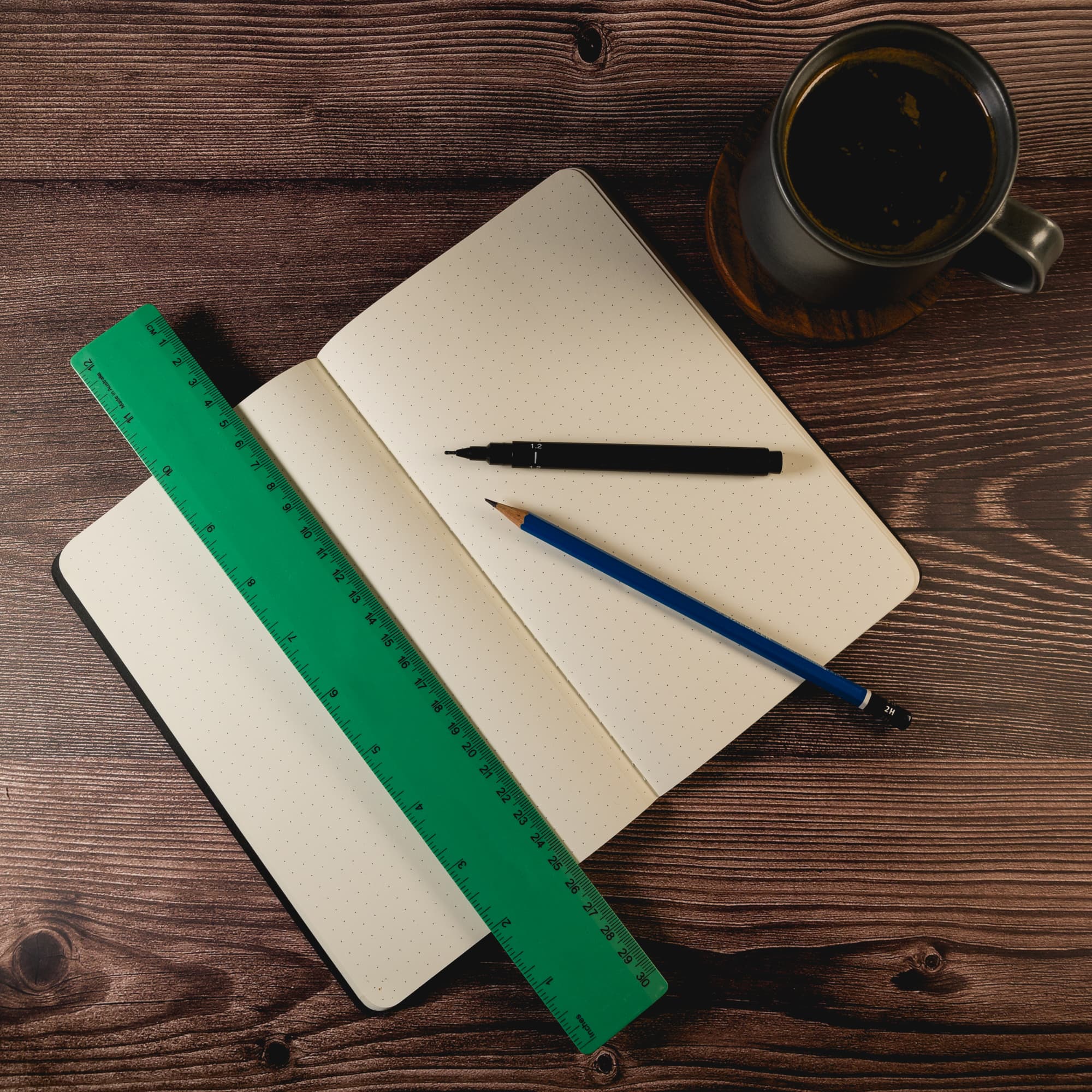 A green 30cm recycled plastic ruler rests beside an open dotted notebook, a black pen, and a blue pencil, with a coffee cup nearby.