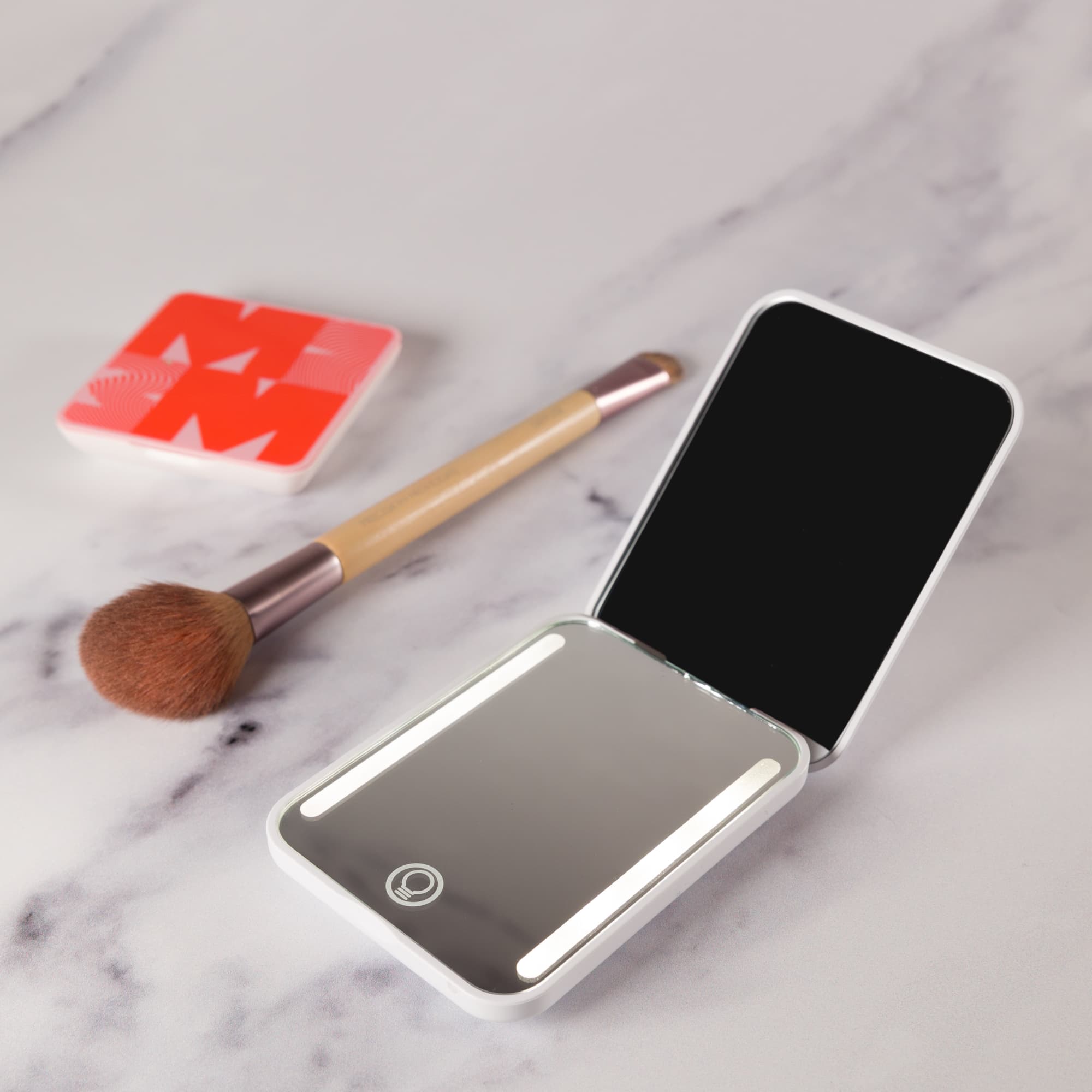 A compact mirror with built-in LED lights, featuring a white casing, alongside a blush compact and brush.