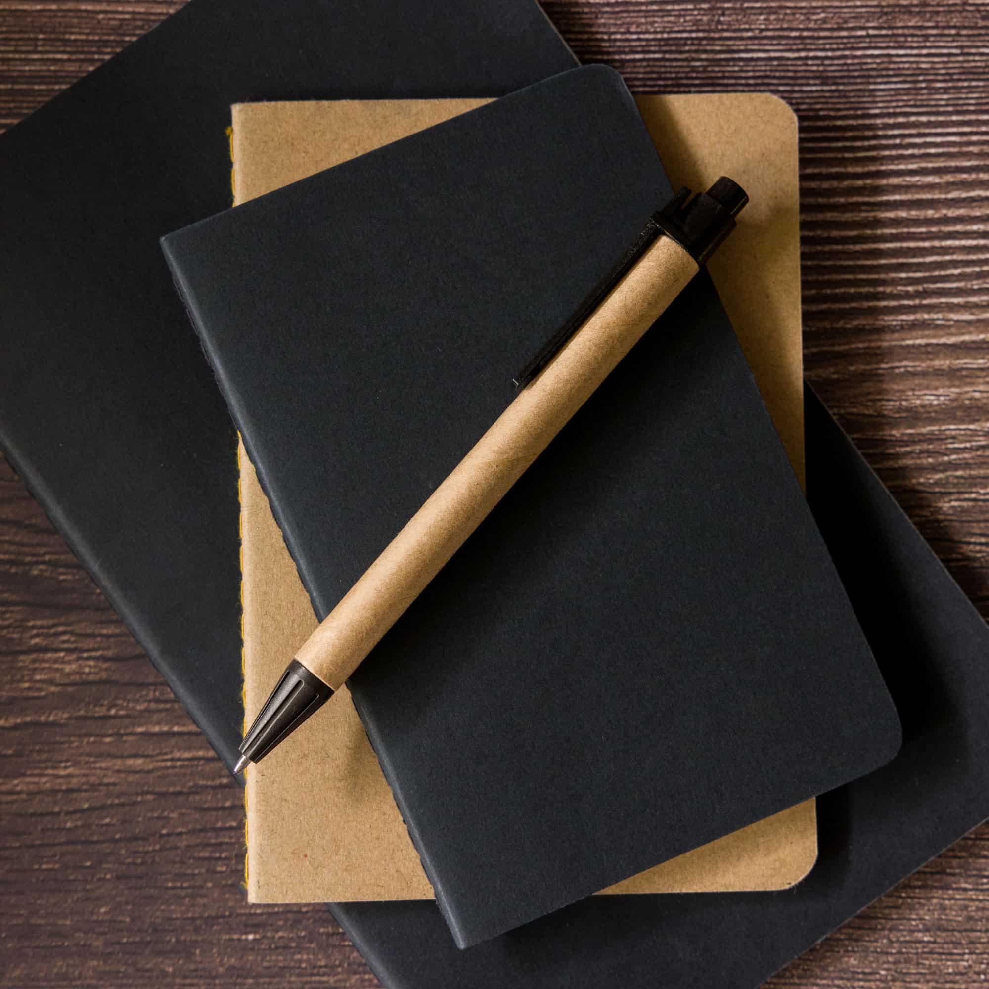 A cardboard pen rests atop two notebooks, one black and one tan, on a wooden surface.