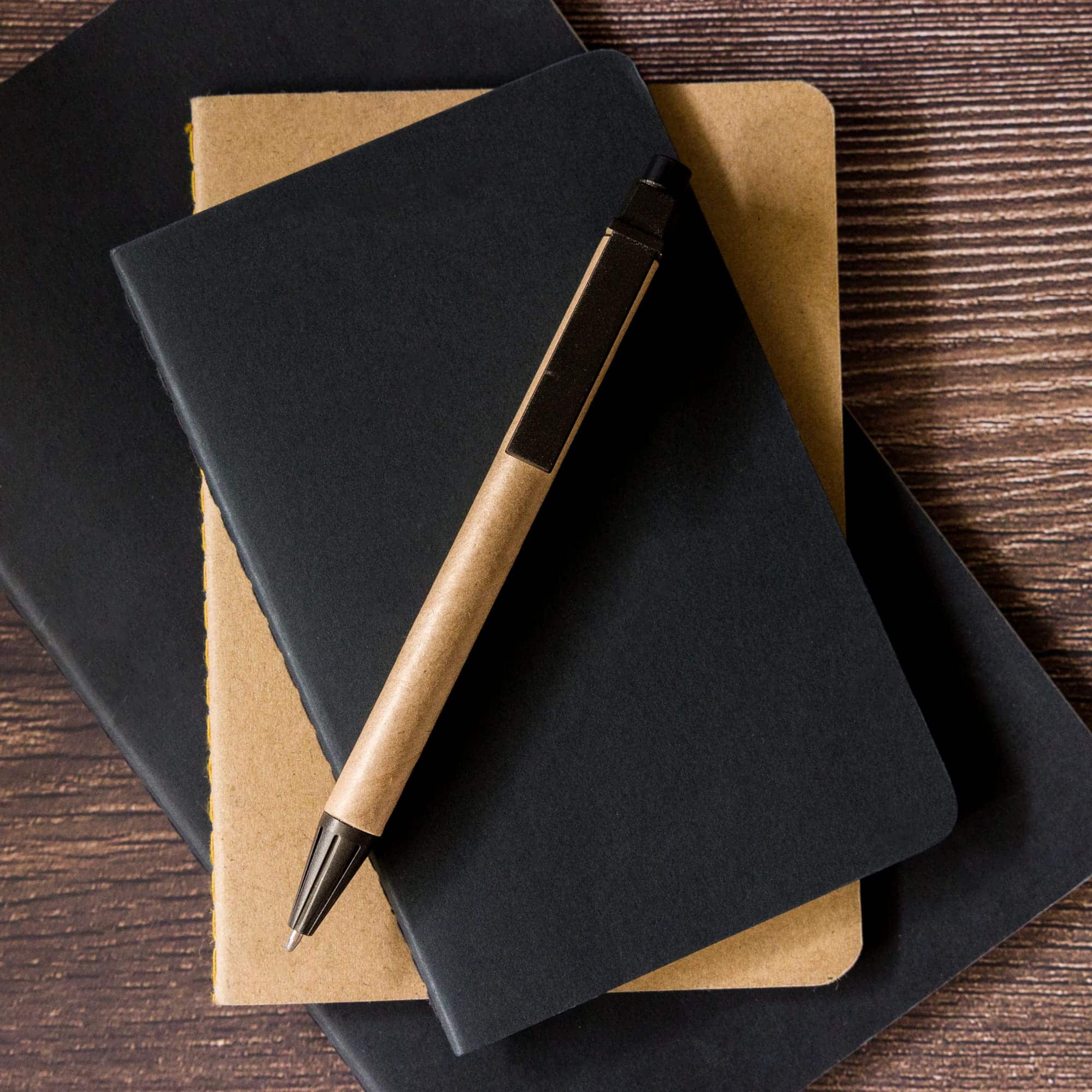 A cardboard pen lies atop two notebooks, one brown and one black, showcasing a sleek and eco-friendly design.