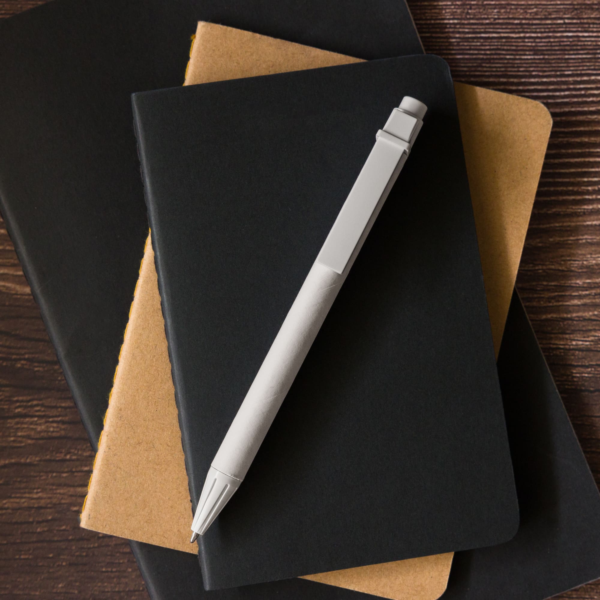 A Matador Cardboard Pen rests on top of two notebooks, one black and one brown.