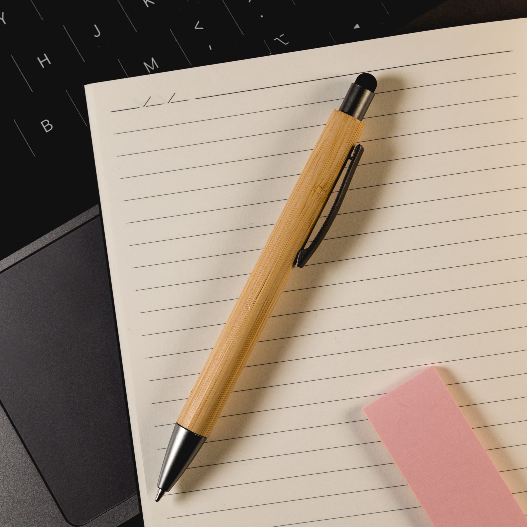 A bamboo pen/stylus with a black tip and clip, positioned on a lined notepad next to a pink sticky note.