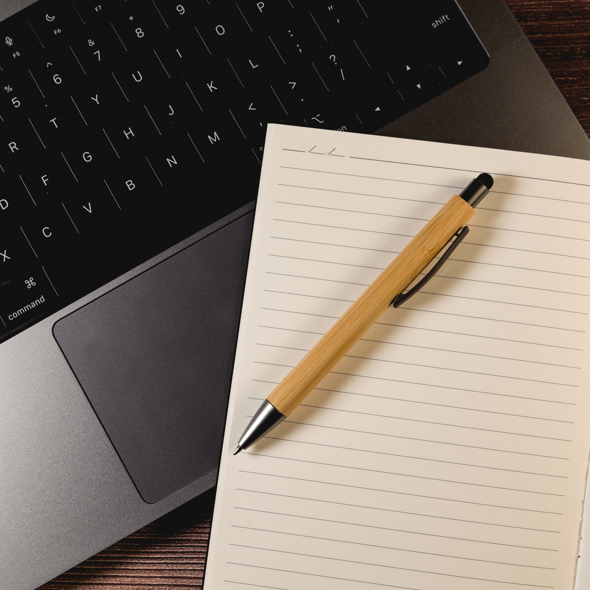 A bamboo stylus pen rests atop a blank notepad, next to a laptop keyboard.