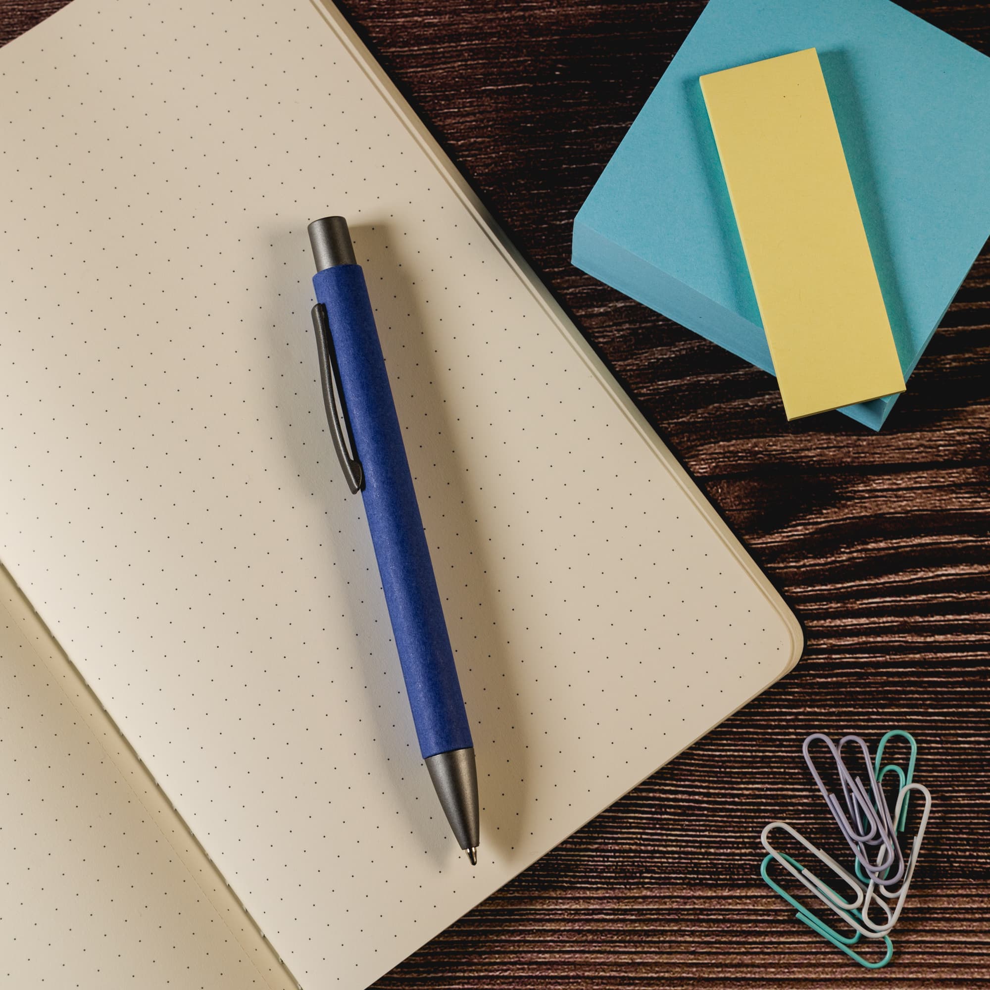 A blue pen placed on a dotted notebook, accompanied by sticky notes and paperclips.