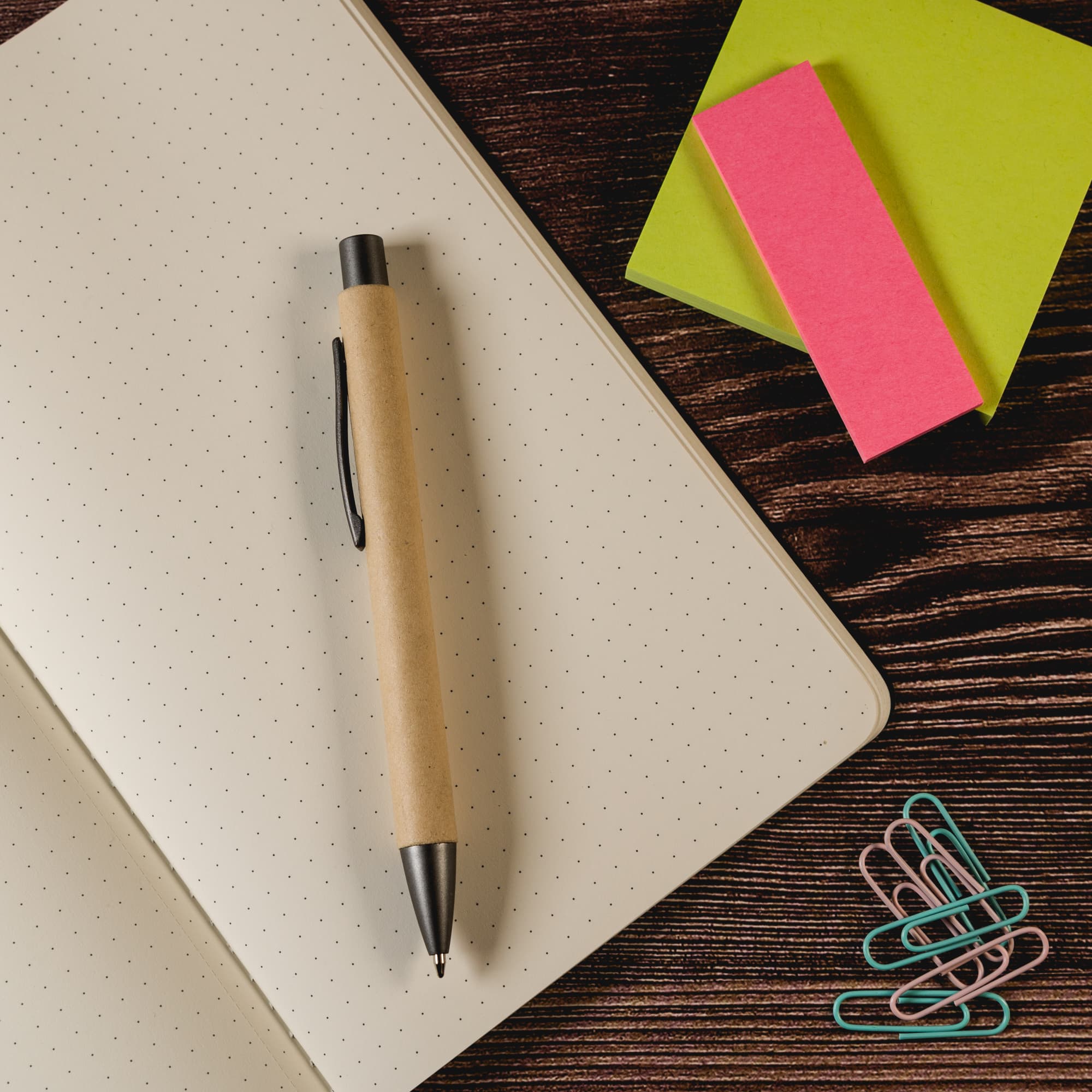 A pen made from kraft paper lies on a dotted notebook, accompanied by colorful sticky notes and paper clips.
