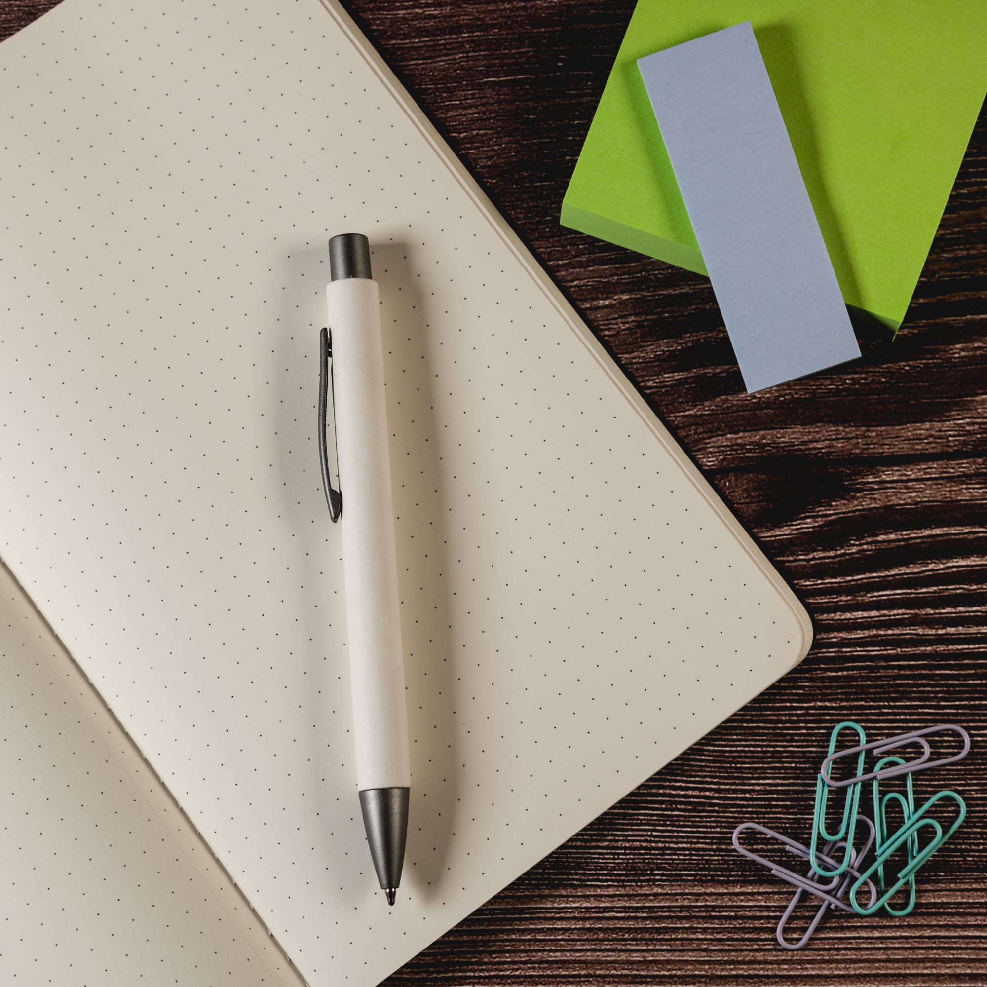 An Aspen Paper Pen rests on a dotted notebook, accompanied by colorful sticky notes and paper clips.