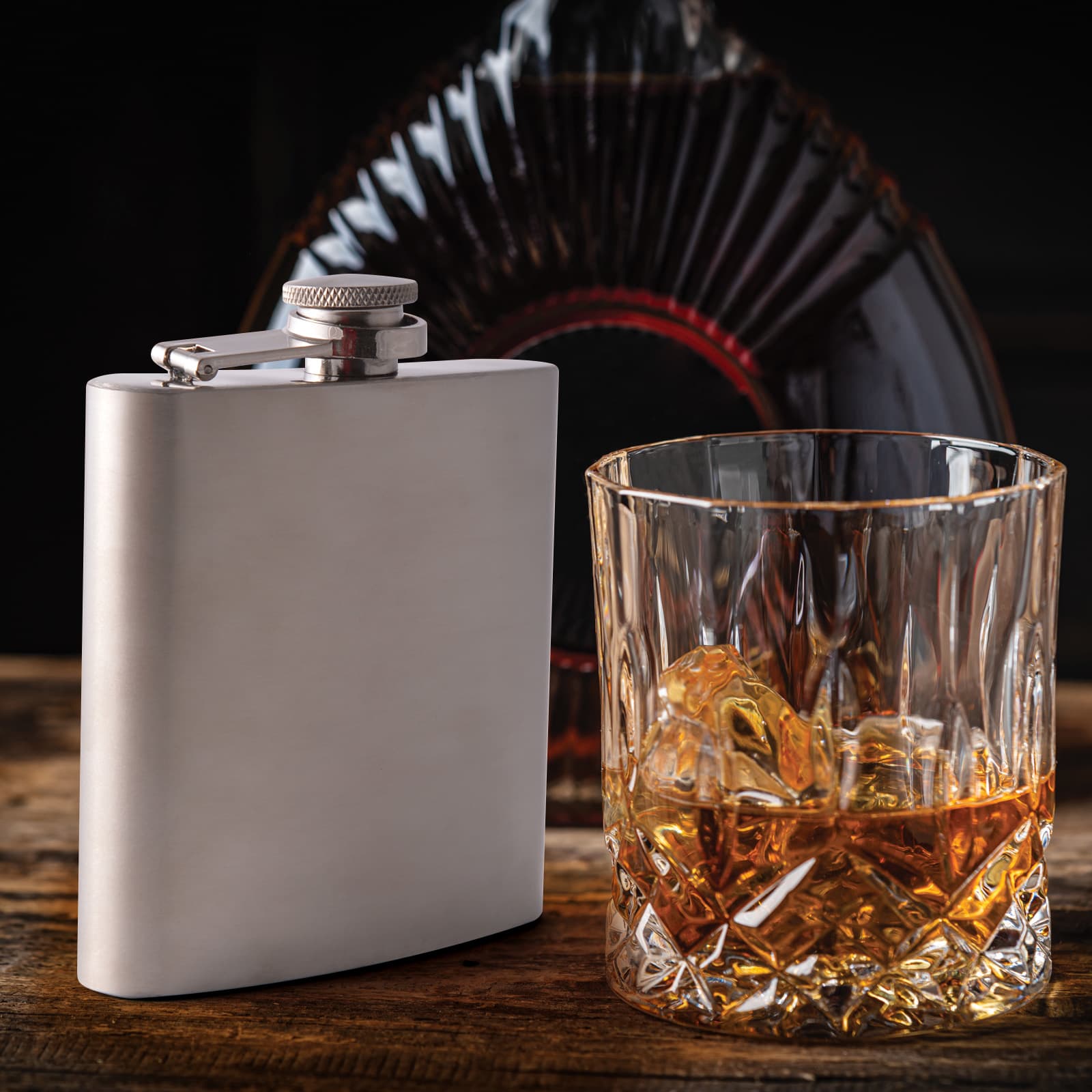A stainless steel hip flask beside a glass filled with amber liquid and ice, set against a dark background.