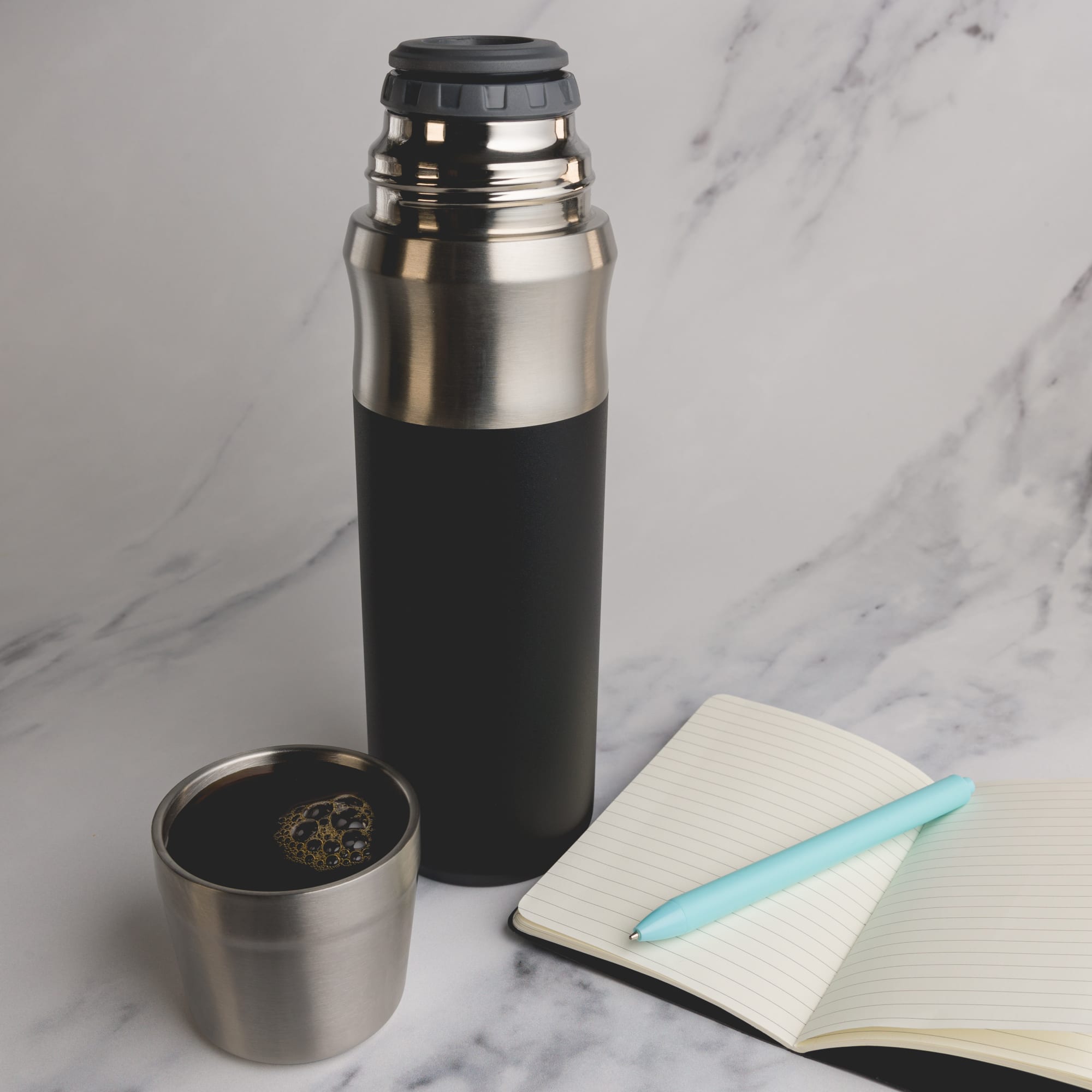 A drink bottle featuring a sleek stainless steel design with a black matte finish, accompanied by a small cup and a notebook.