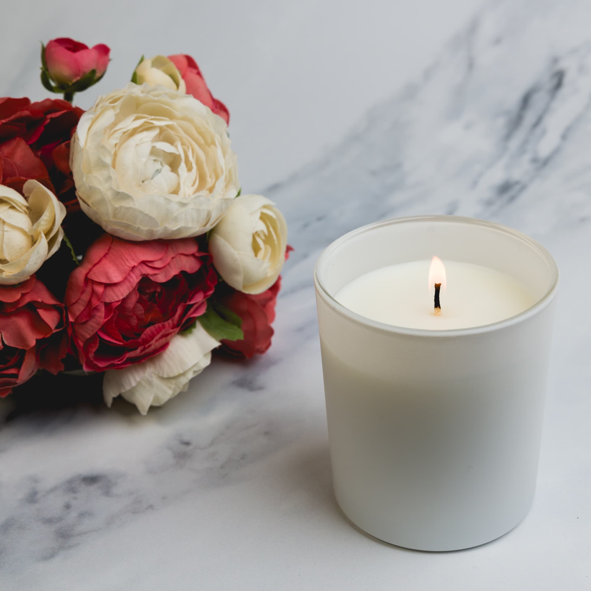 A large Gleam Glass Candle in a white holder with a lit wick sits next to a bouquet of mixed-colored flowers.