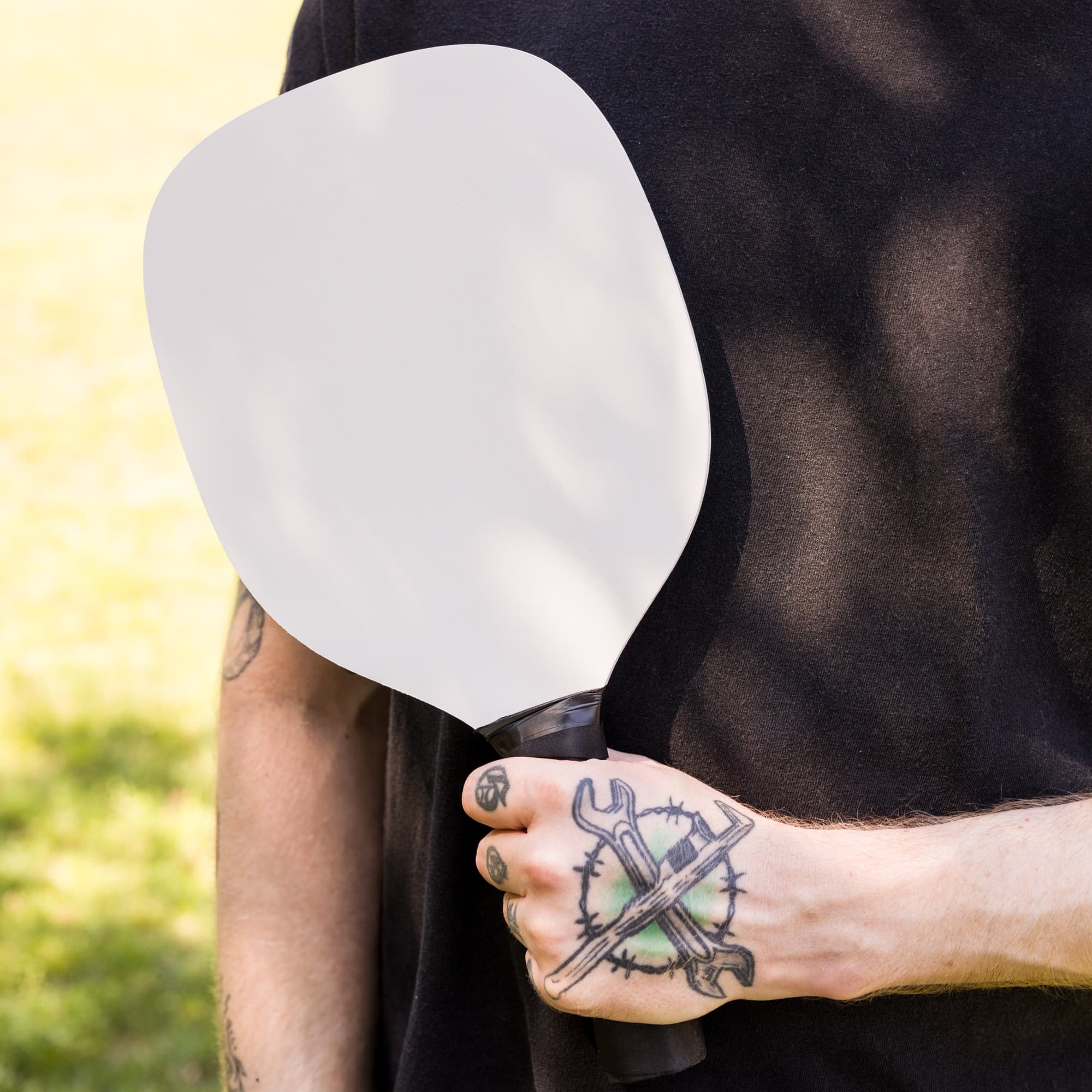 A pickleball paddle with a blank surface, held by a person with a tattooed hand.