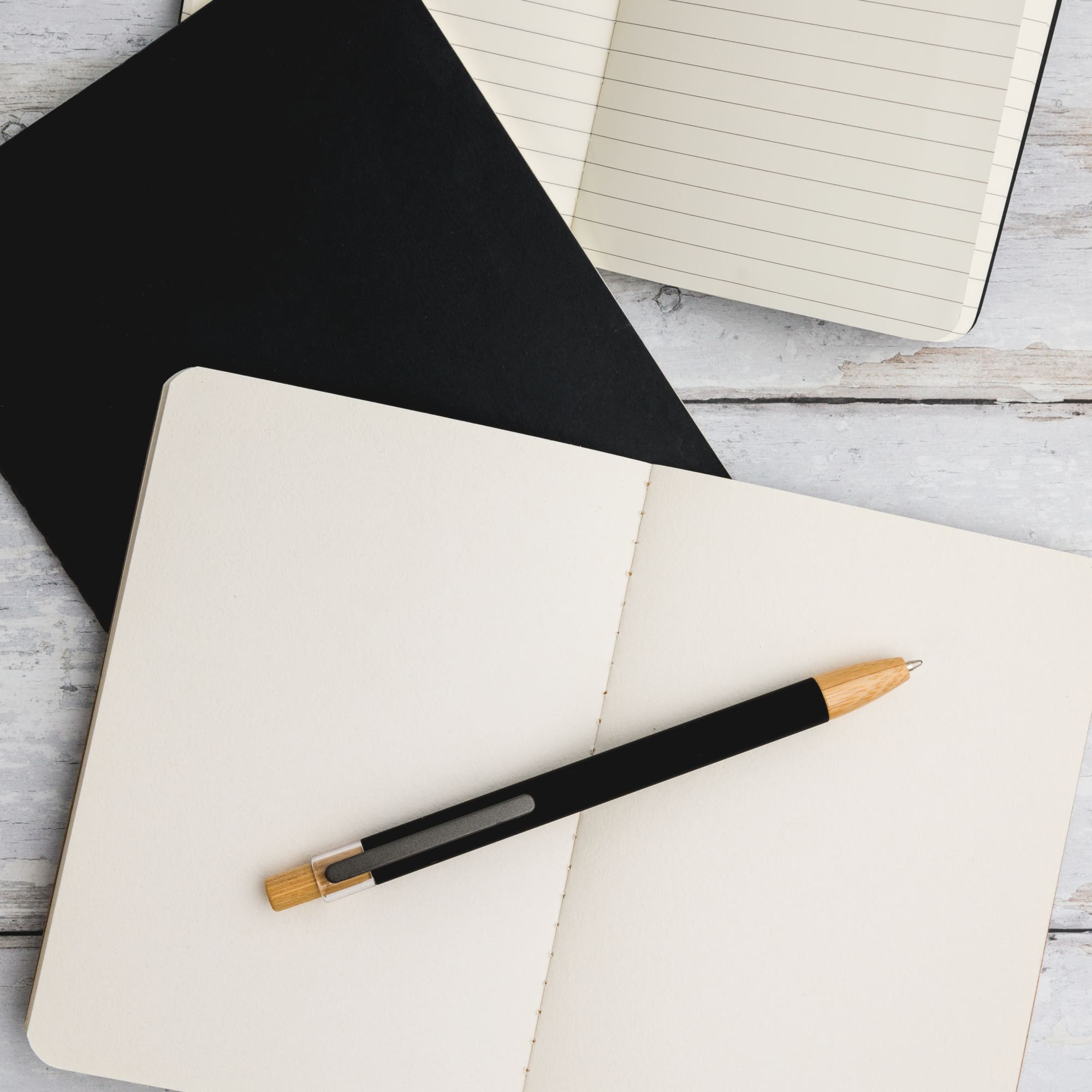 A black aluminium pen with a bamboo accent rests on an open cream-colored notebook, featuring a black notebook above it.