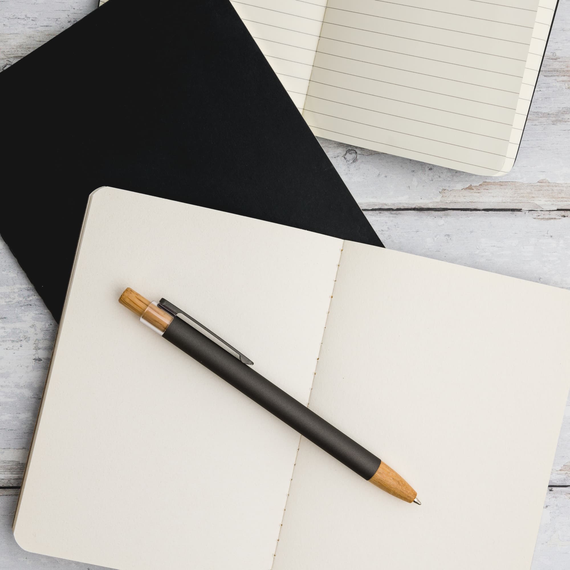 A Serenity Aluminium Pen rests on an open blank notebook, accompanied by a lined notebook and a black notebook.