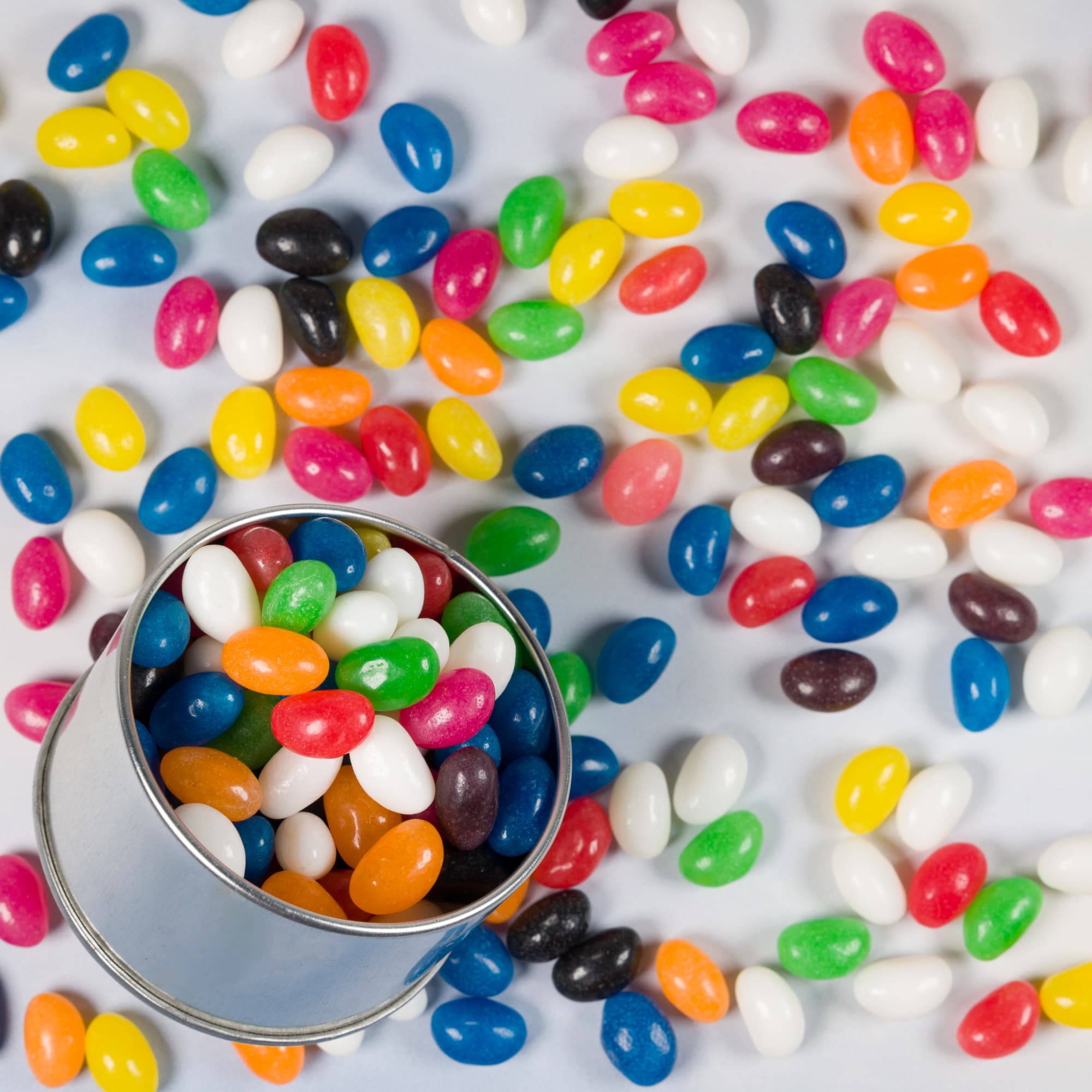 A silver round tin filled with assorted colourful jelly beans, surrounded by additional jelly beans on a white surface.