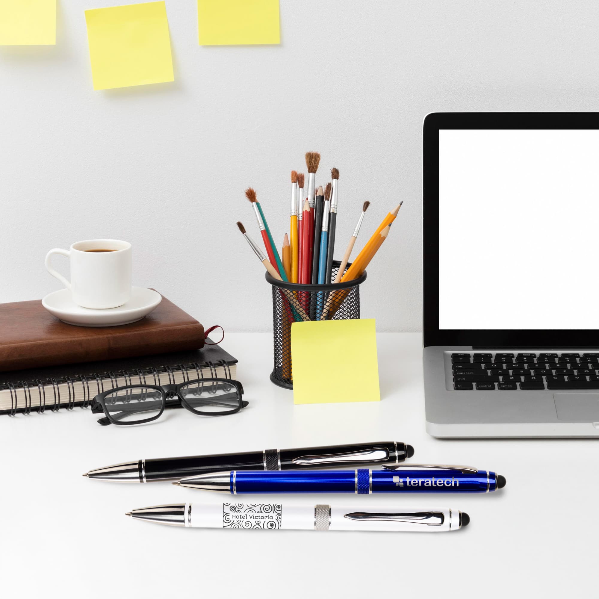 A collection of three metal pens in blue and white, with a coffee cup, notepad, and laptop on a desk.
