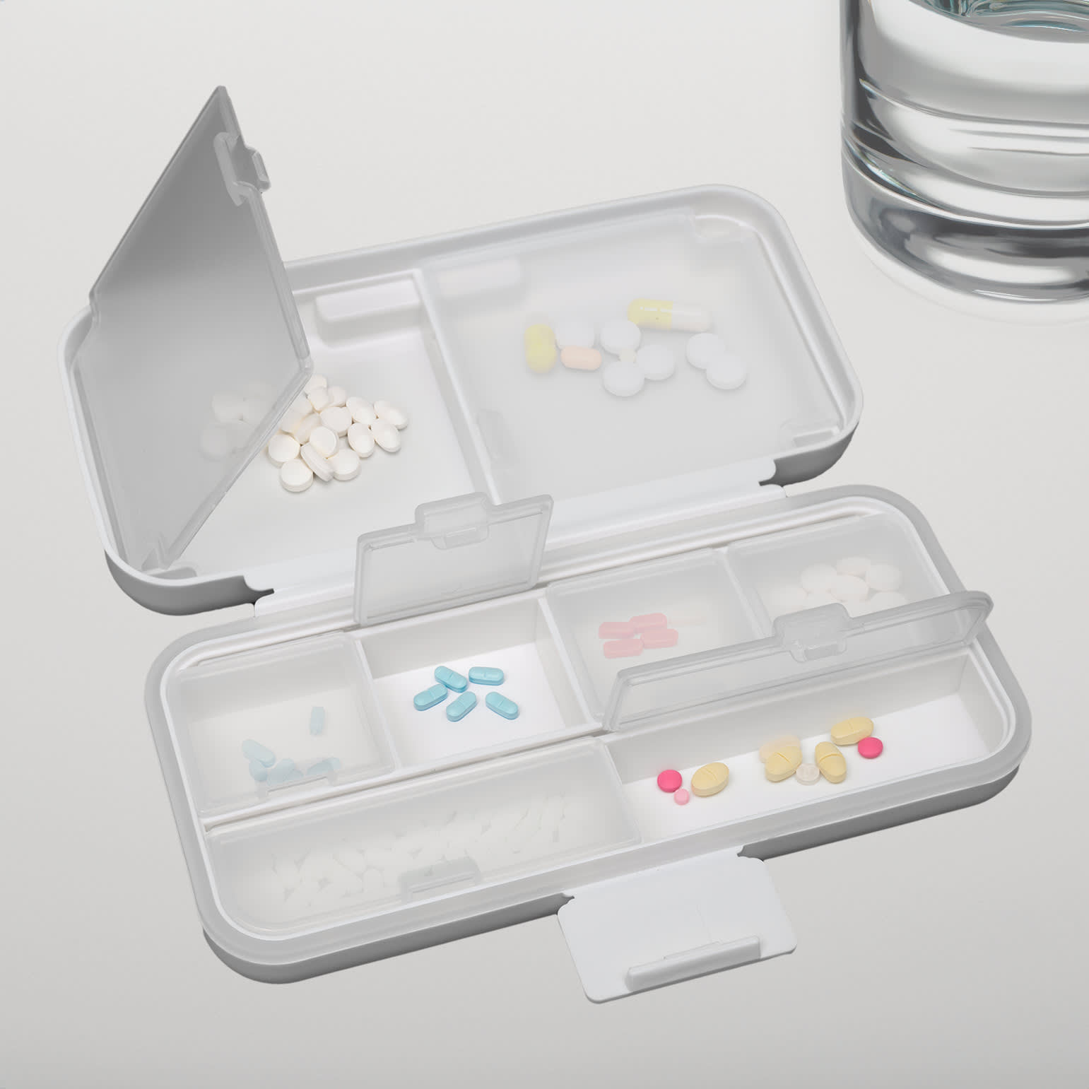 A compact white pill organizer with multiple compartments, filled with various colored tablets, beside a glass of water.