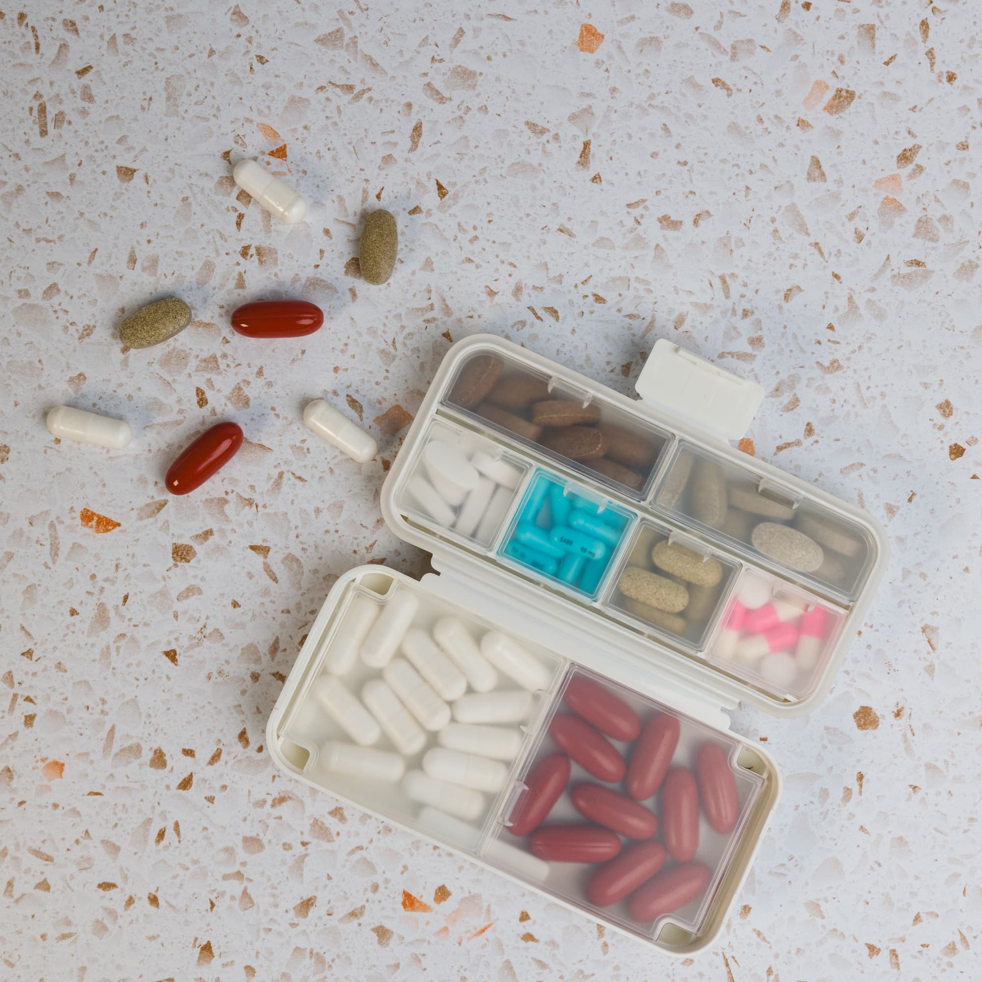 A compact white pill organizer with multiple compartments filled with various colored pills scattered around.
