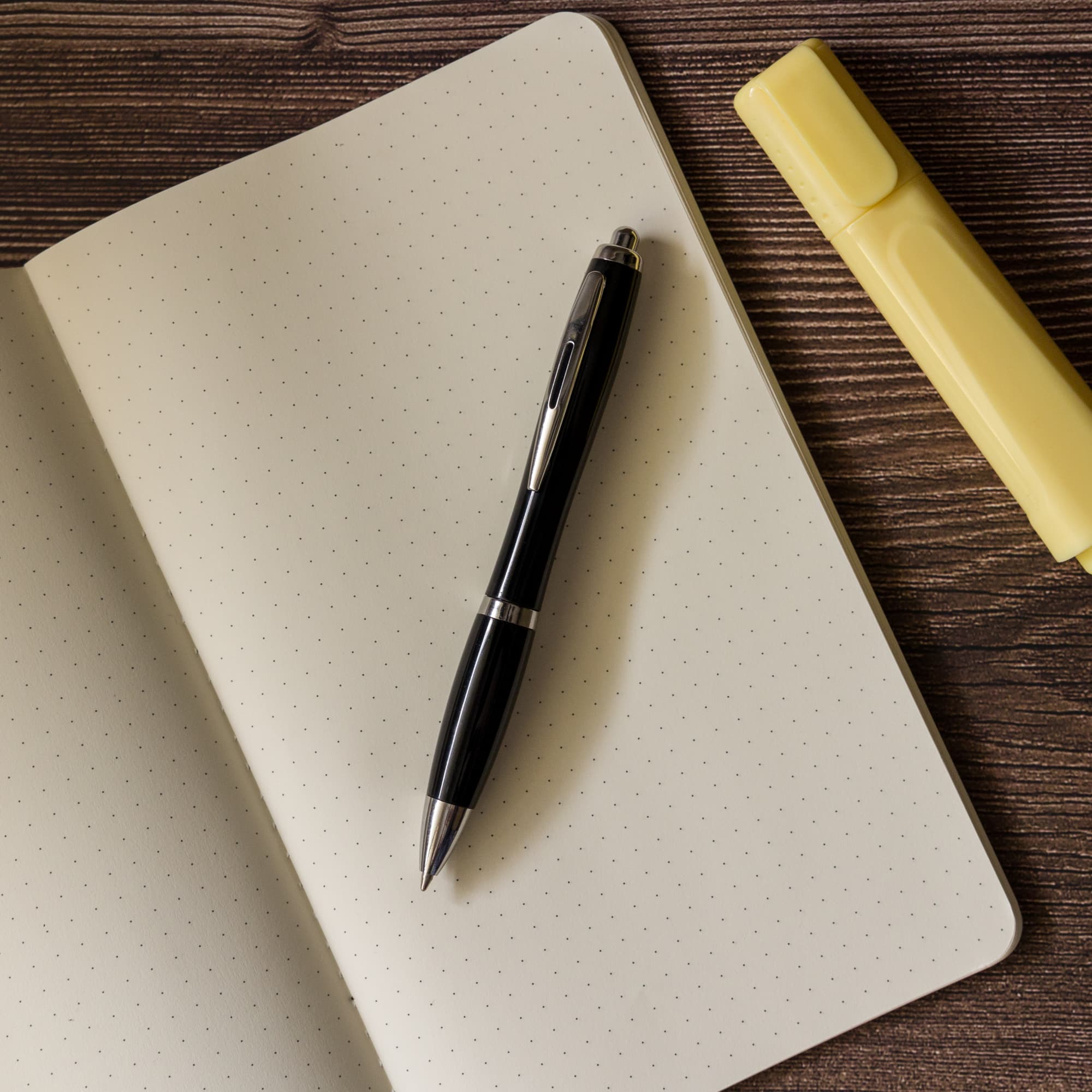 A metal pen is resting on a dotted notebook, accompanied by a yellow highlighter.
