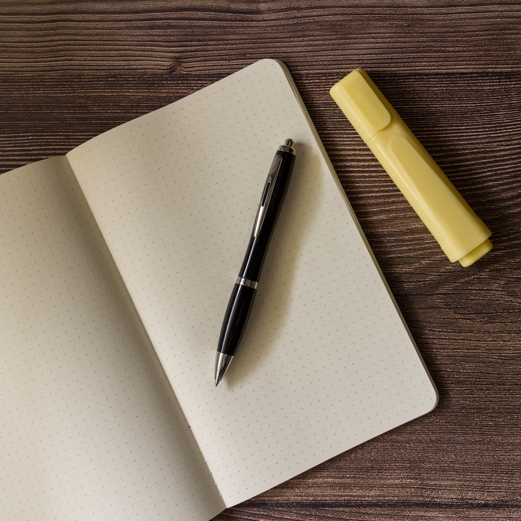 A metal pen rests on an open notebook with dotted pages, alongside a yellow highlighter.