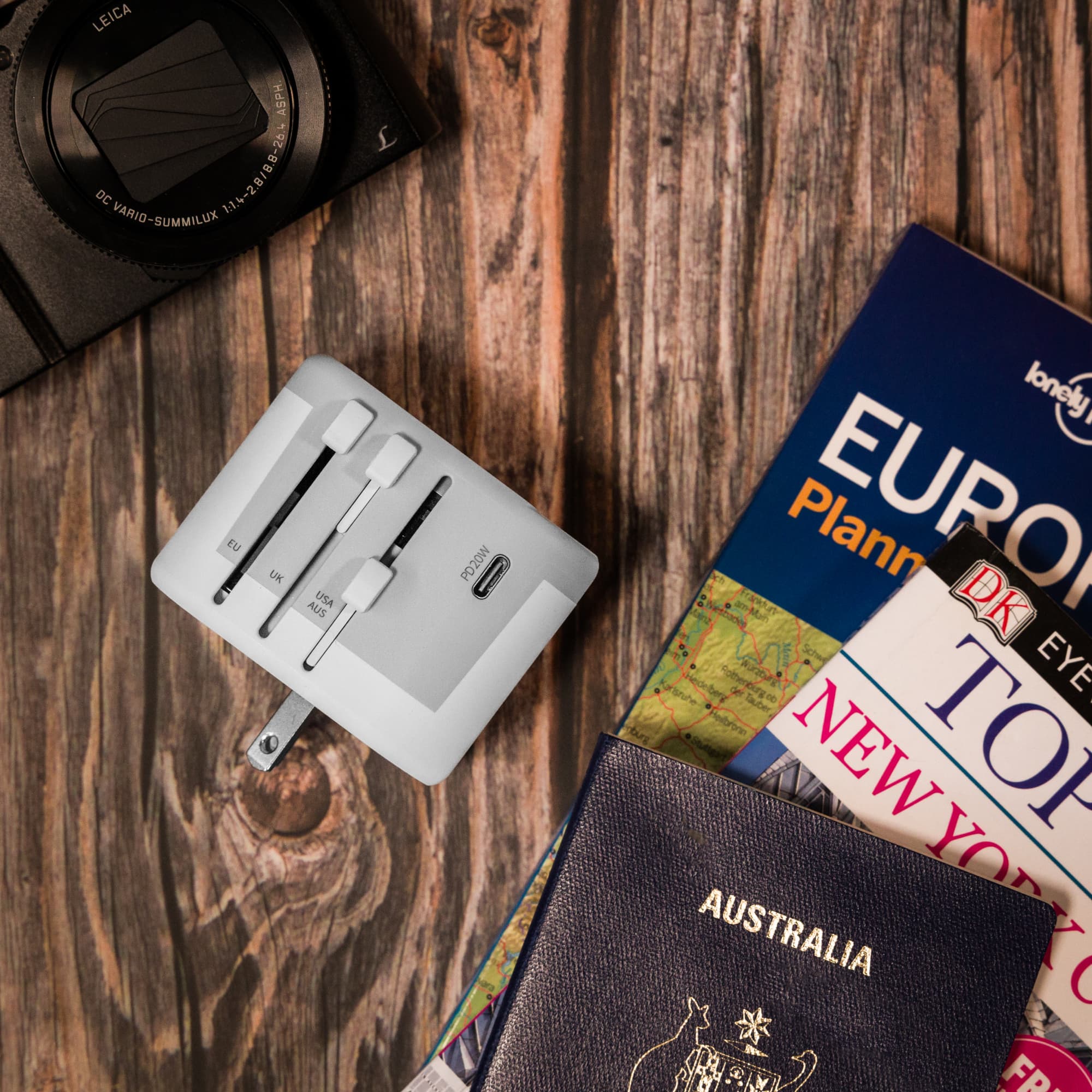 A white travel adaptor rests on a wooden surface beside travel guides, a camera, and a passport.