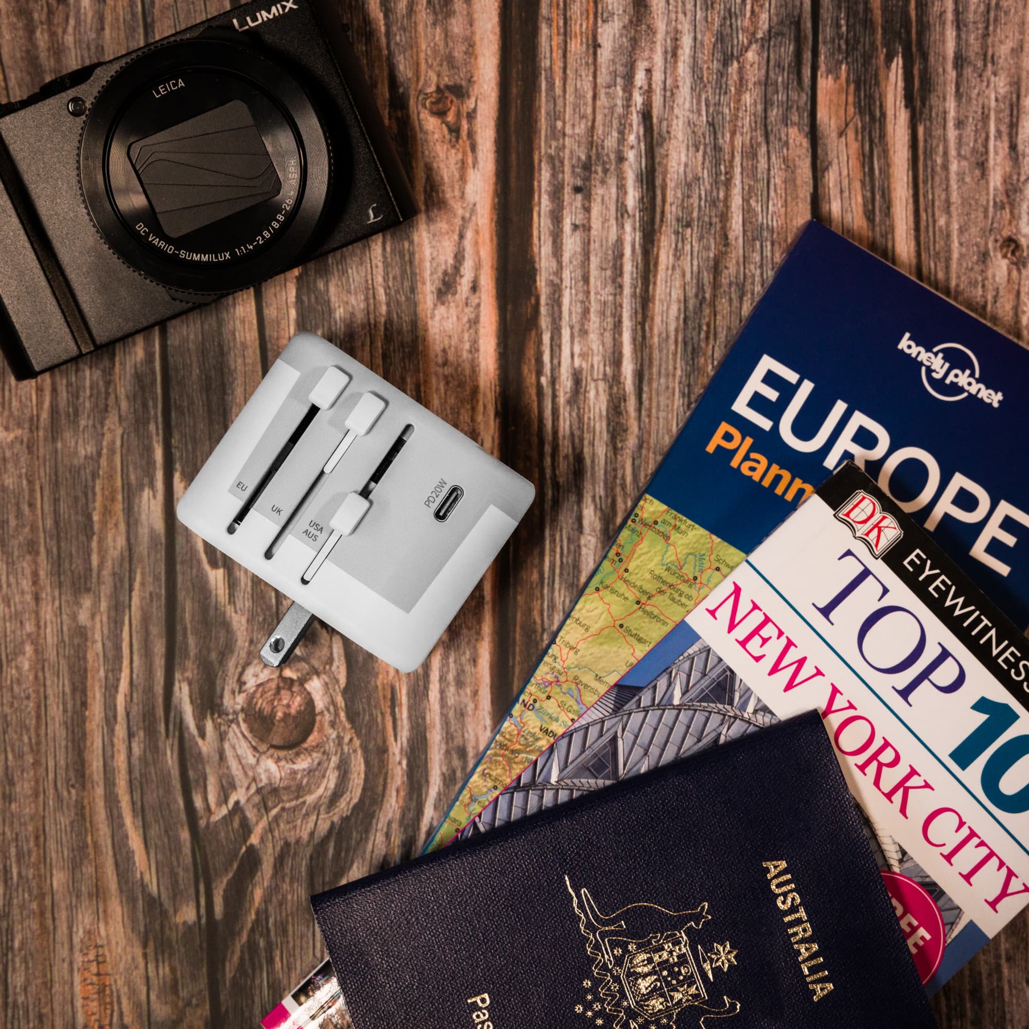 A travel adaptor positioned on a wooden surface alongside travel books, a passport, and a camera.