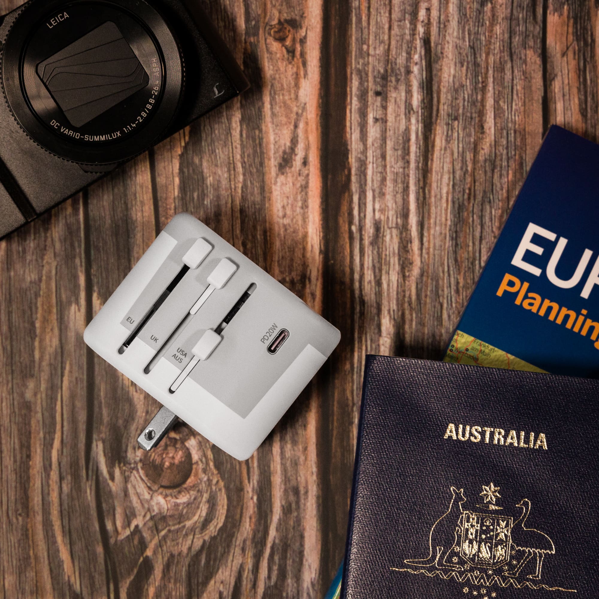 A travel adaptor placed on a wooden surface alongside a camera, travel guide, and a passport.