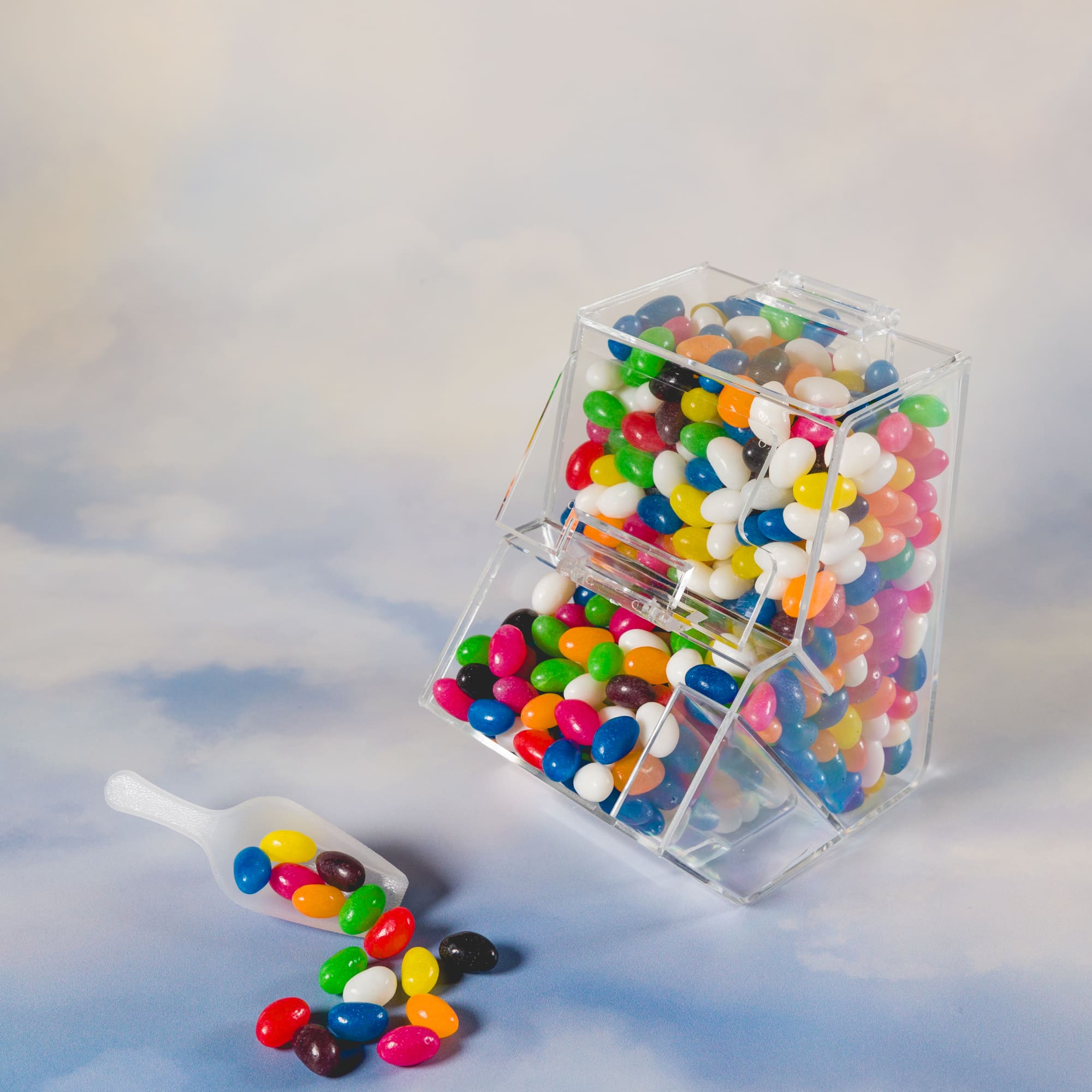 A clear dispenser filled with assorted color mini jelly beans, with a scoop resting nearby.