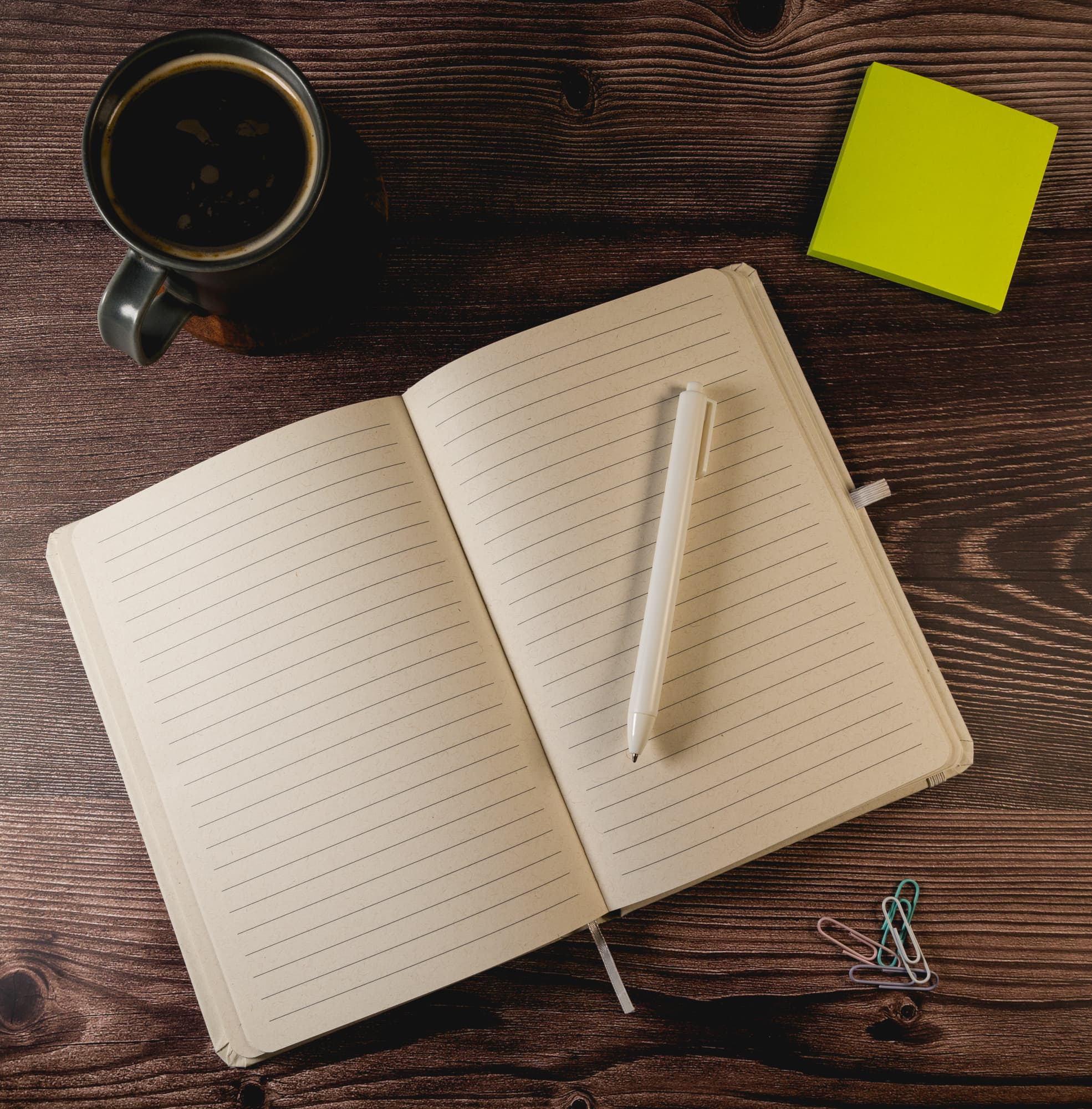 An open A5 notebook with lined pages, a white pen, a green sticky note, and paper clips on a wooden surface.