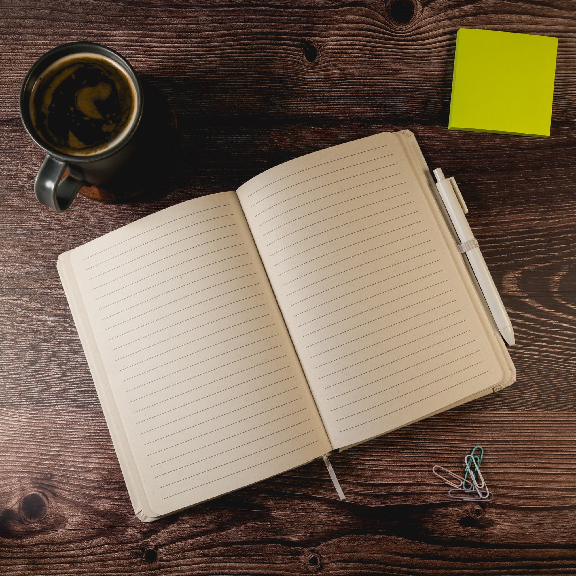 An open beige notebook with lined pages, a pen, paper clips, a green sticky note, and a cup of coffee on a wooden surface.