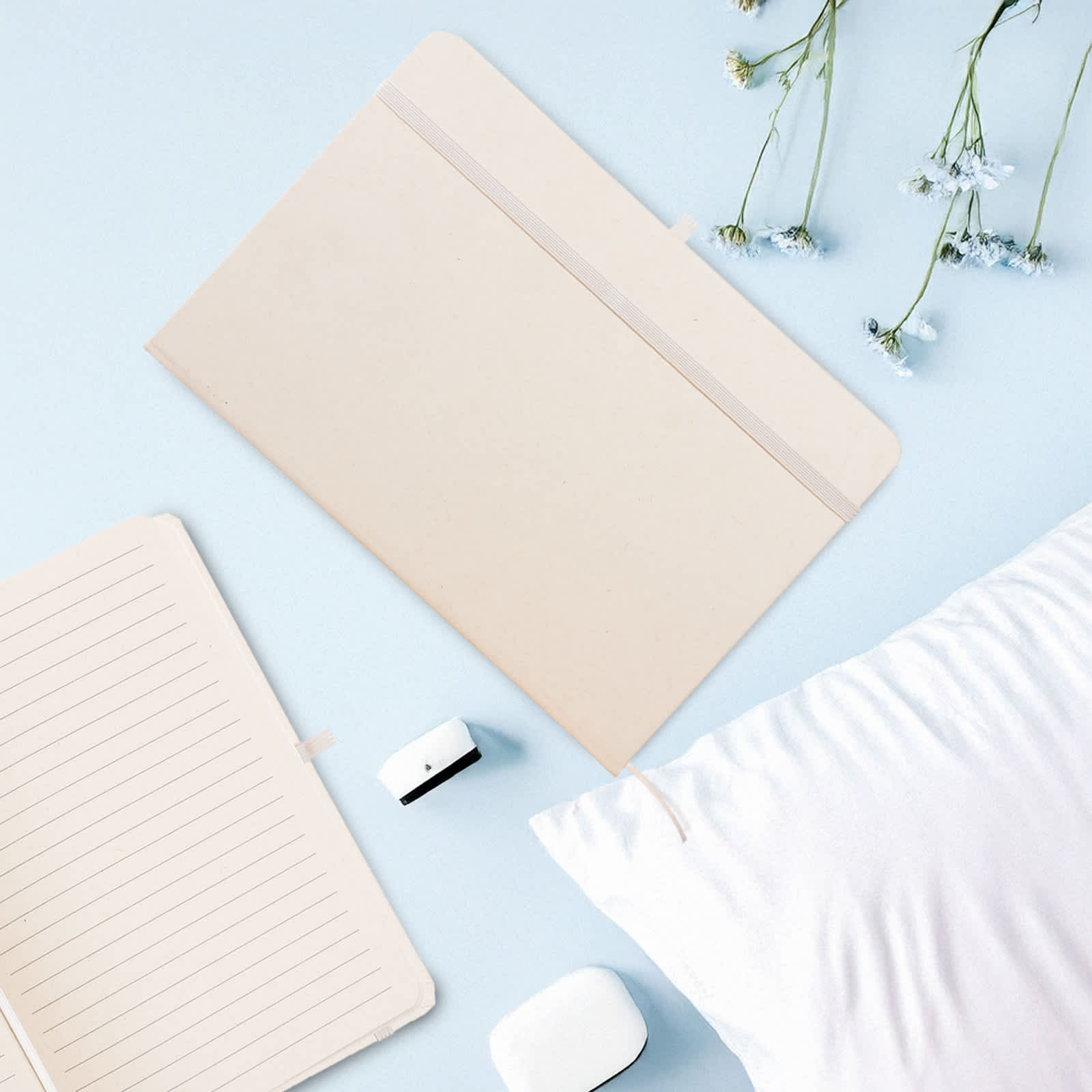 A beige A5 notebook with lined pages, surrounded by a white pillow and small white flowers. It has a logo.