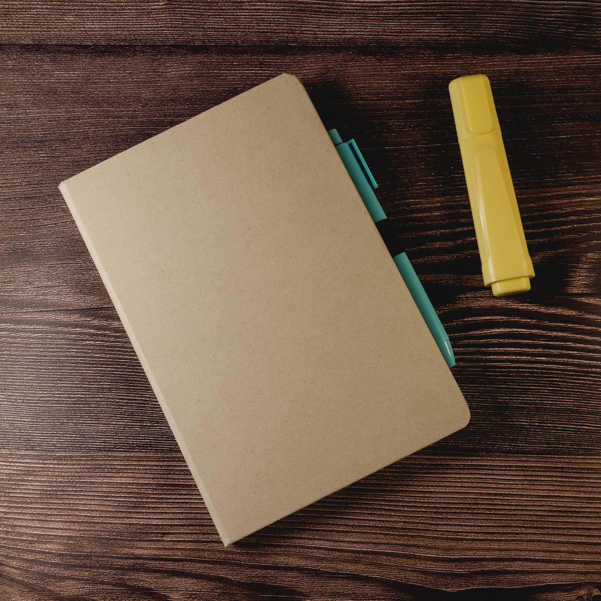 A Venture Sugarcane A5 Notebook accompanied by a blue pen and a yellow highlighter, set on a wooden background.