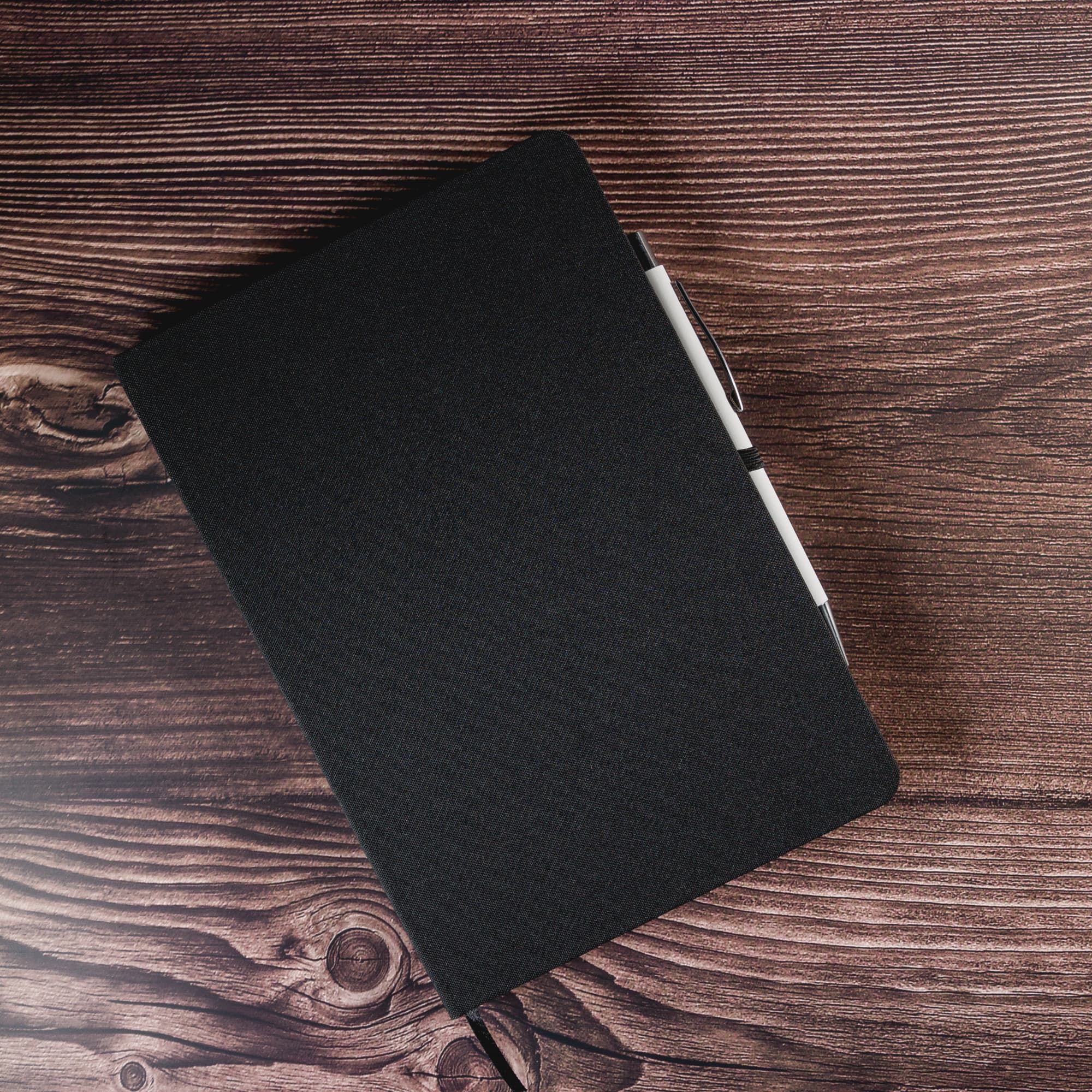 A black A5 notebook with a textured cover and an attached pen on a wooden surface. It features a logo on the front.
