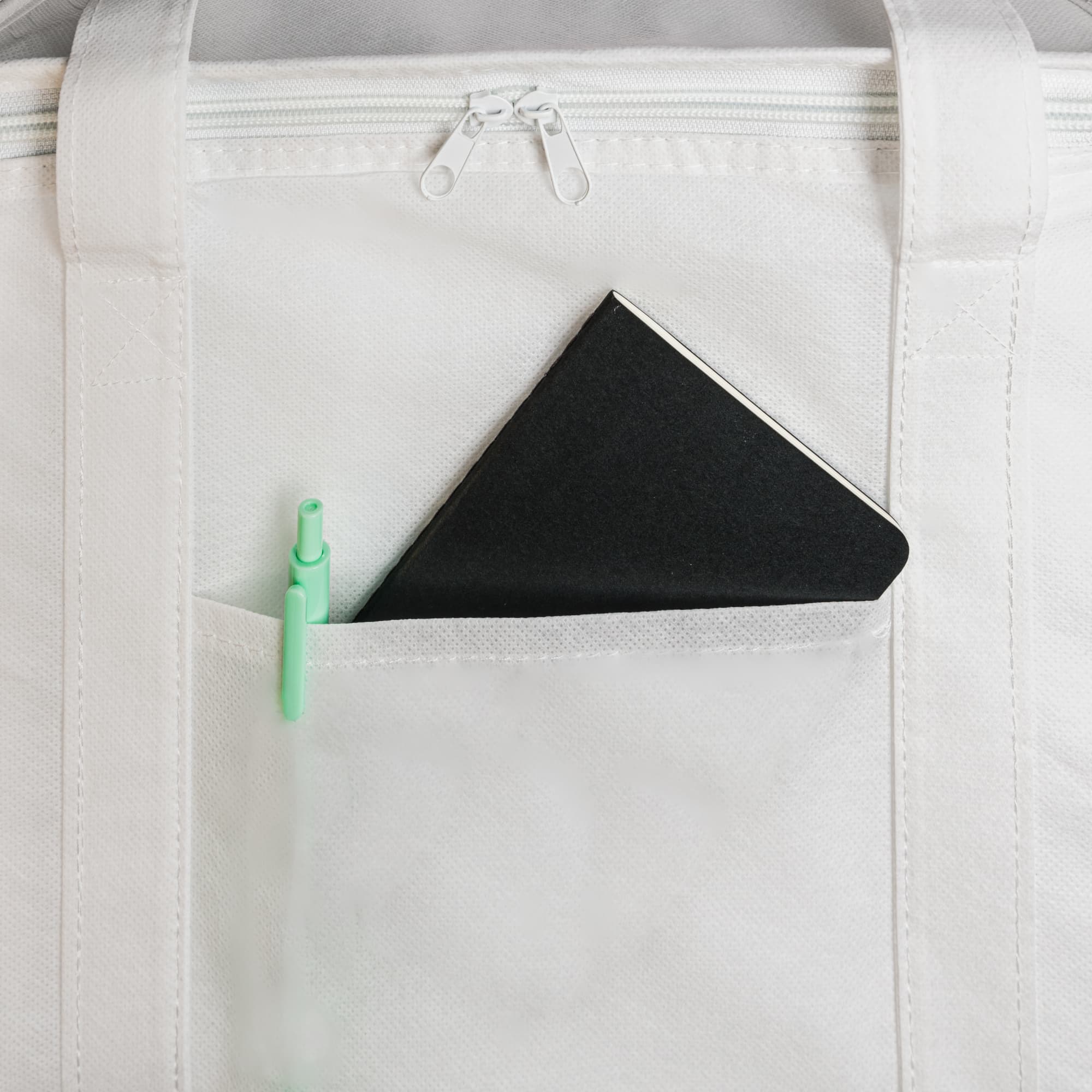 A black notebook and green pen are visible in a front pocket of a white cooler bag. The bag features a zippered closure.