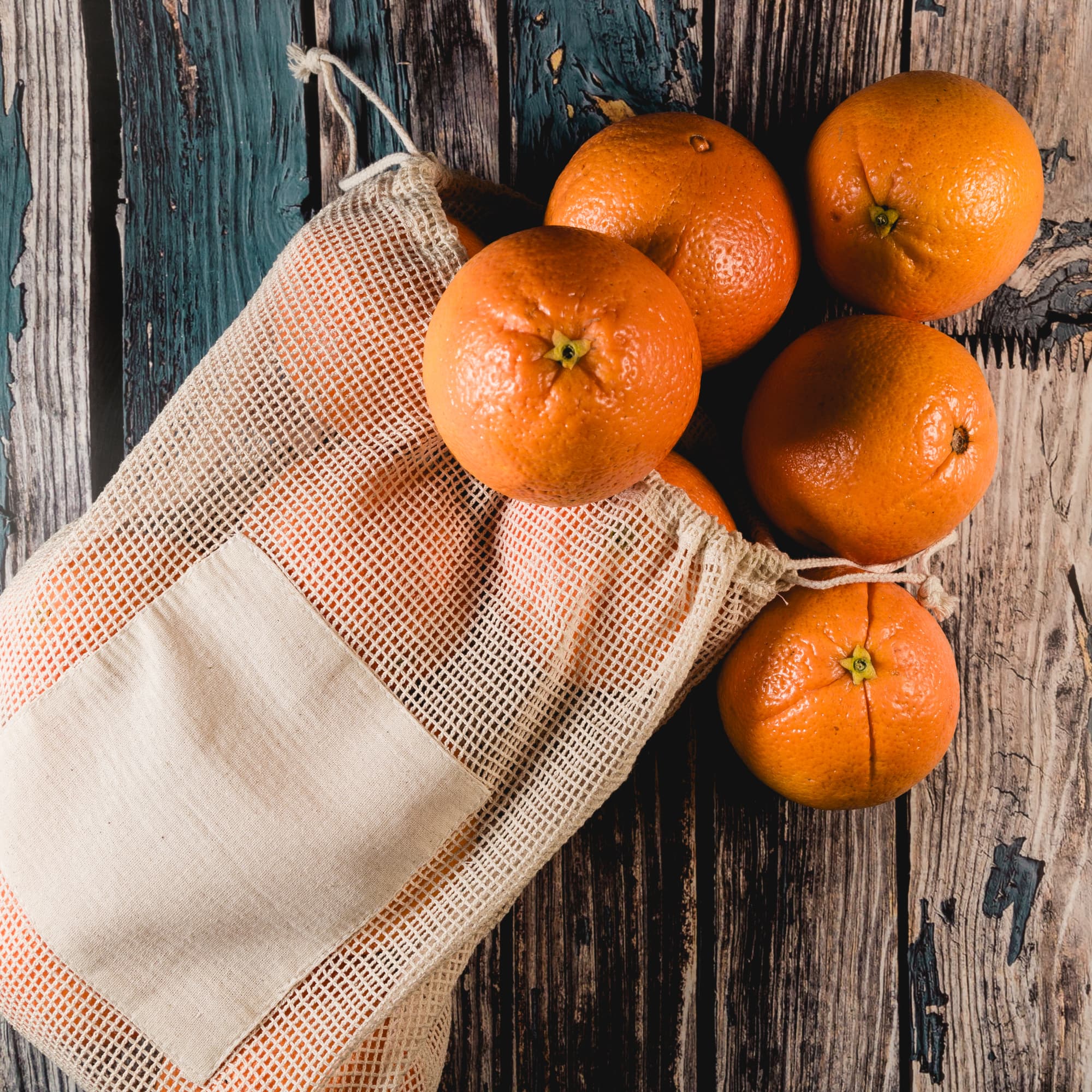 A mesh produce bag containing several oranges is placed on a wooden surface alongside additional oranges.