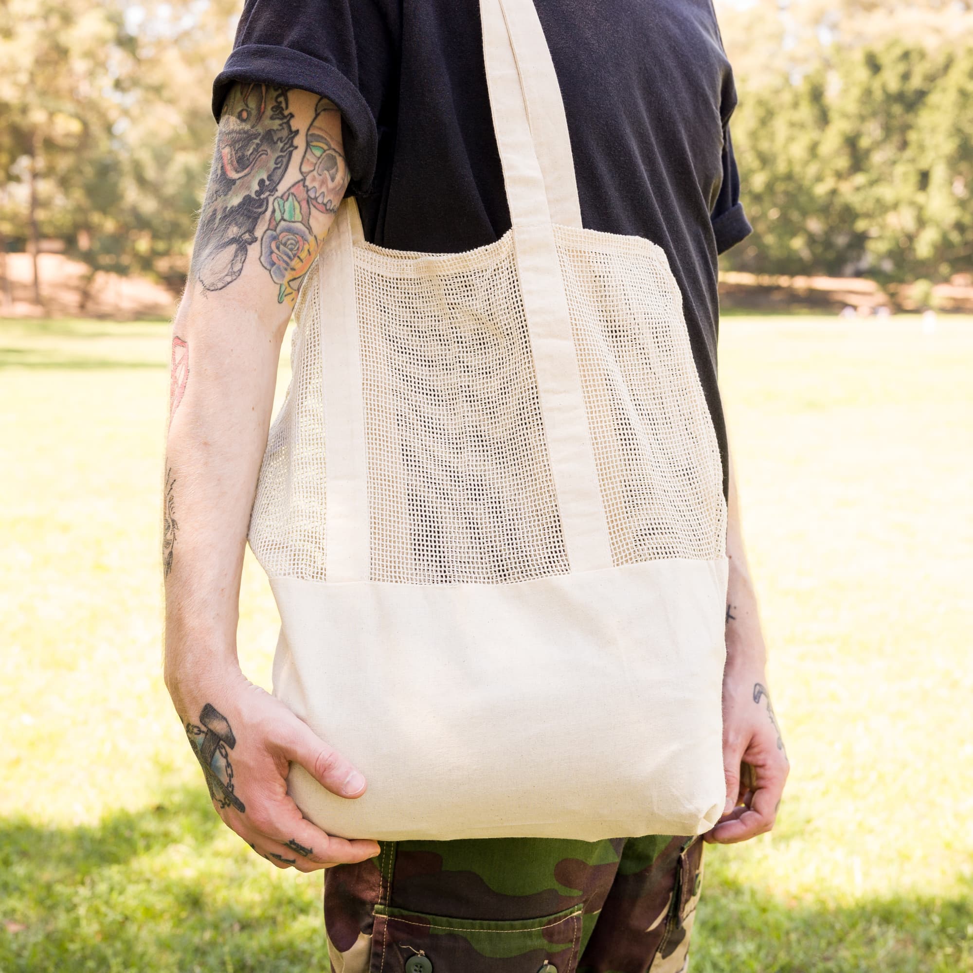 A person holds a beige tote bag with a mesh design, set against a green outdoor backdrop.